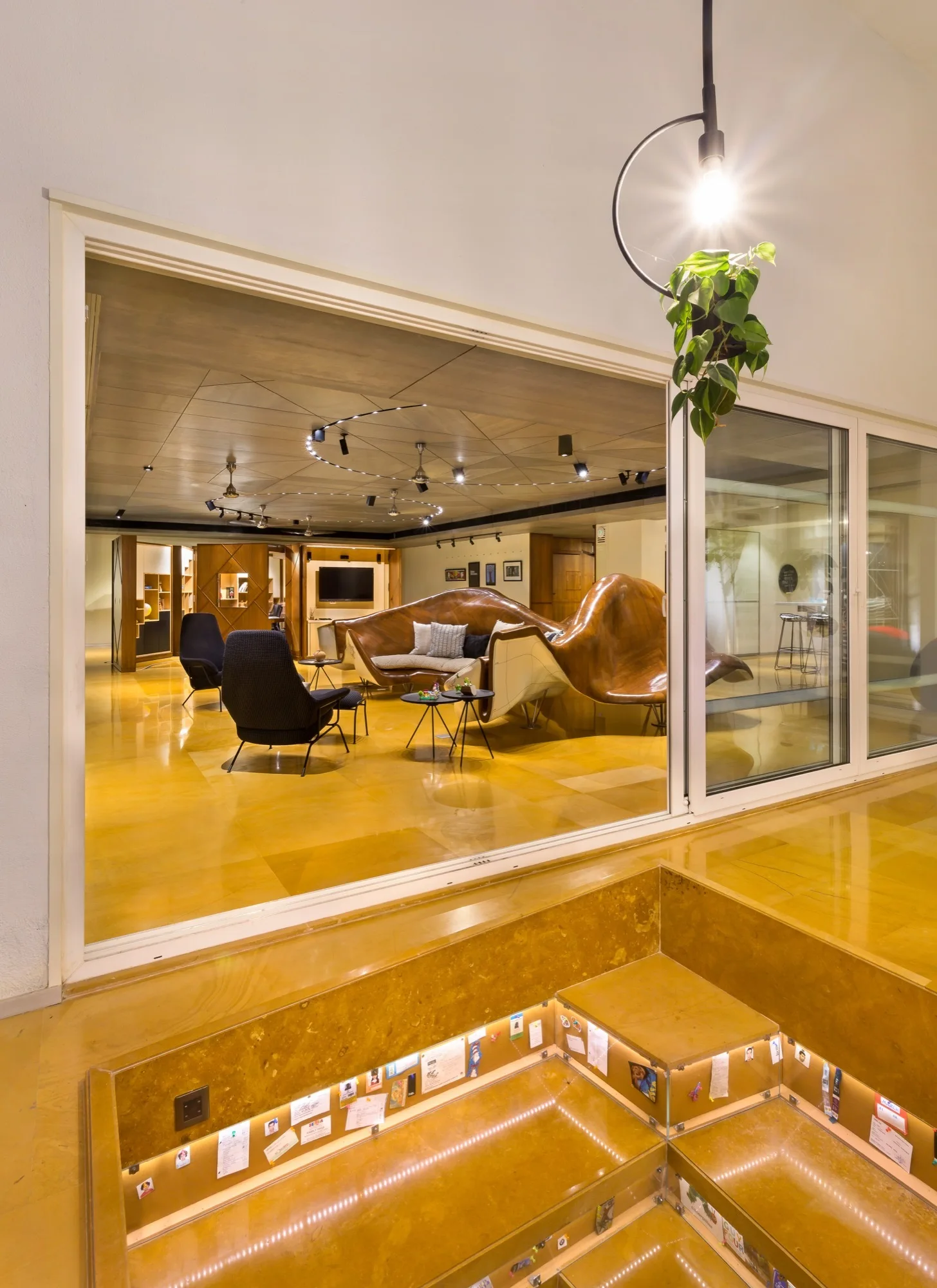 A view through a glass partition into a contemporary lounge with a large sculptural wooden sofa, dark armchairs, and reflective ochre flooring.