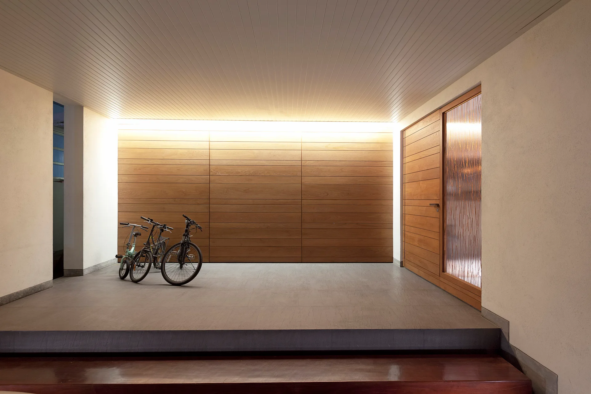 A covered entryway features a horizontal wood paneled wall illuminated by cove lighting, a wood door with textured glass, and parked bicycles.