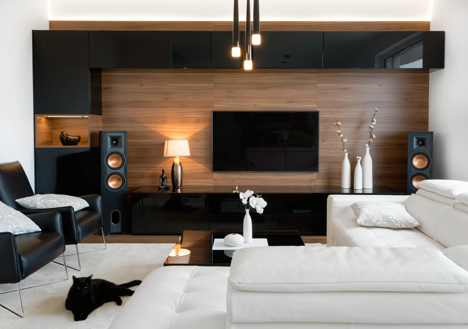 A modern living room features a wood-paneled accent wall, built-in black gloss cabinets, a white leather sectional sofa, and two black armchairs.
