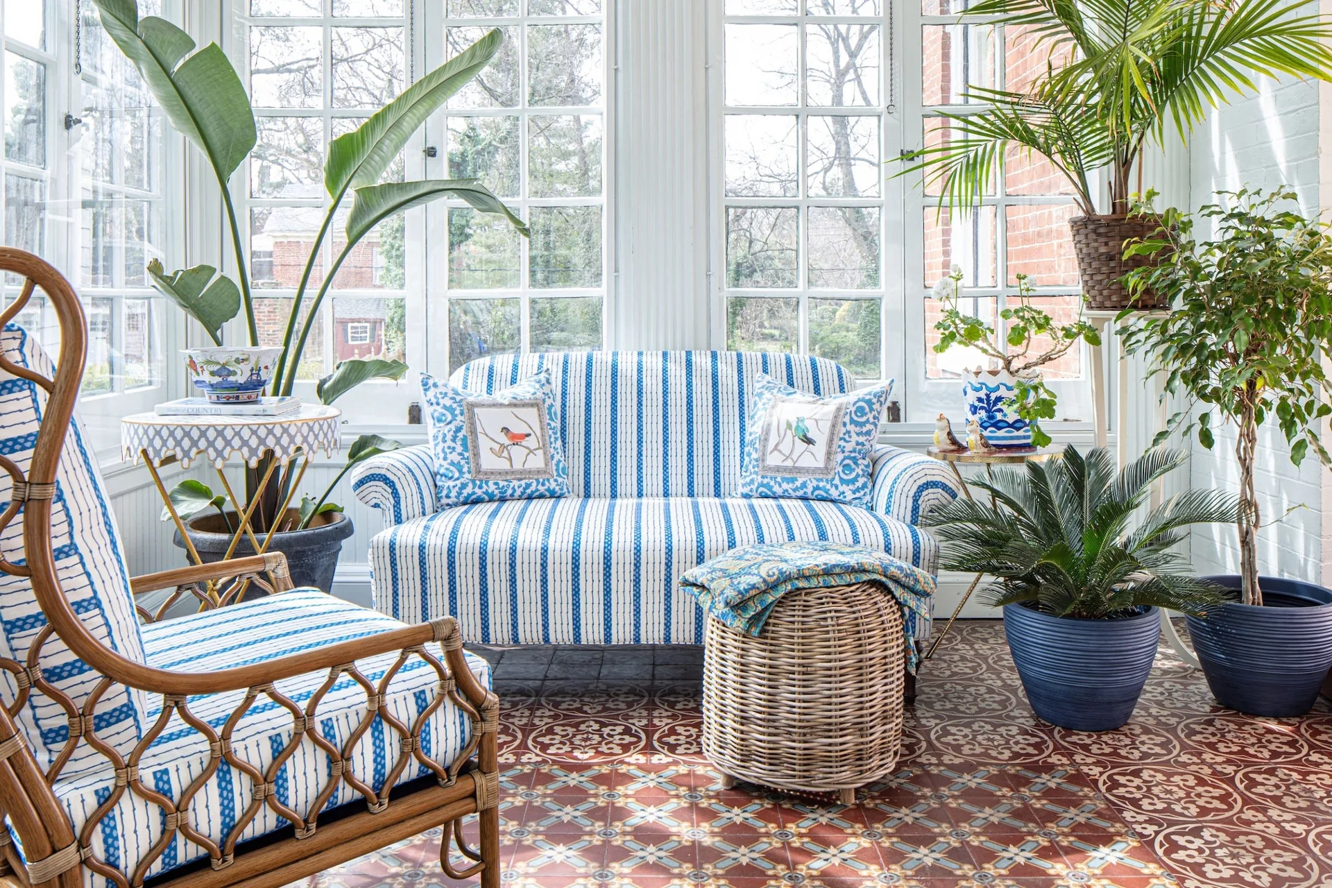 A bright sunroom features a blue and white striped sofa, a rattan armchair, a wicker ottoman, and patterned tile flooring.
