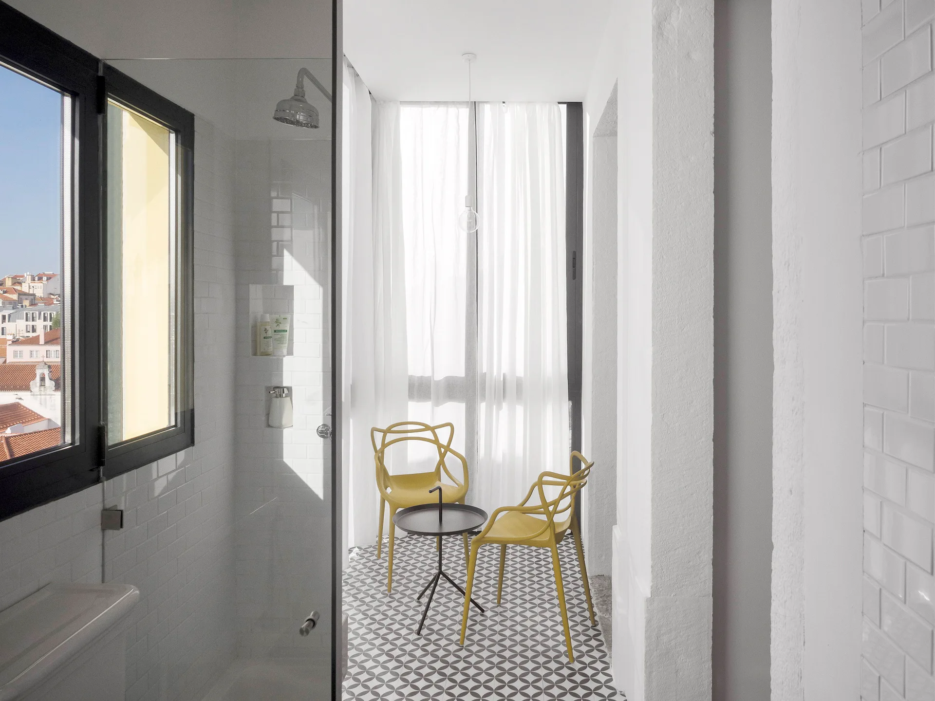 A white tile shower looks out onto a sunlit balcony featuring geometric patterned floor tiles, two yellow plastic chairs, and a black side table.