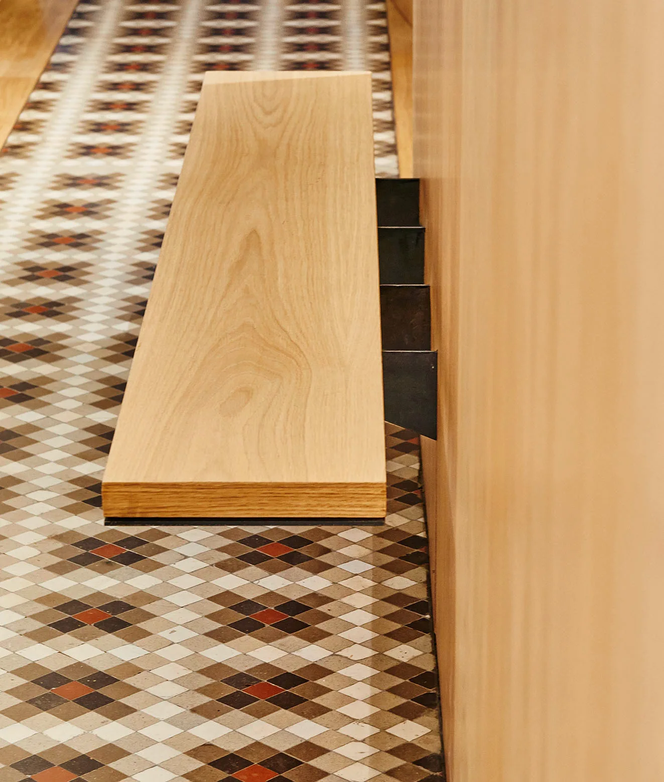 A light wood floating shelf mounted with dark metal brackets onto a wood-paneled wall above a geometric diamond-patterned tile floor.