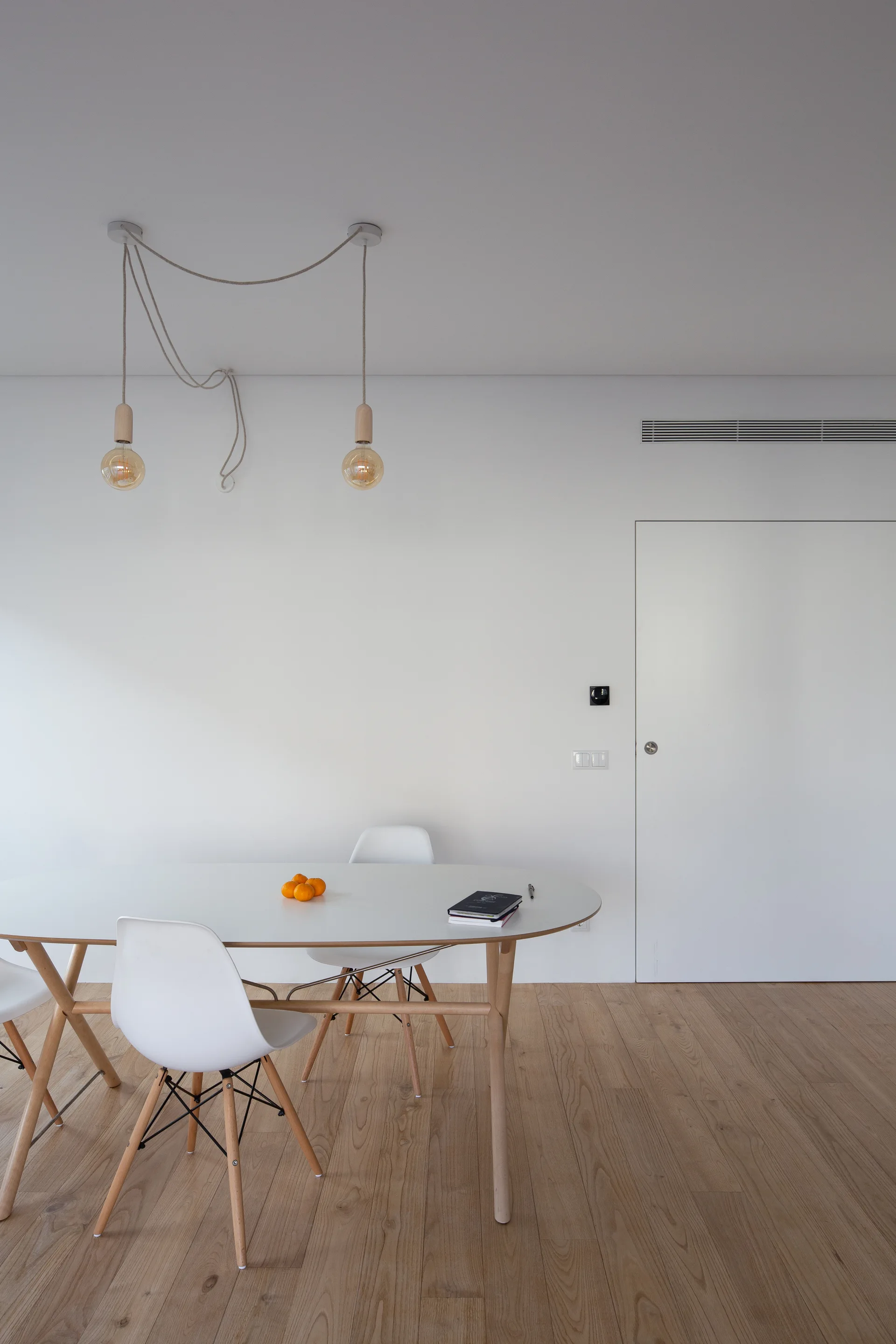 A minimalist dining area with a white oval table, wooden-legged chairs, and two exposed bulb pendant lights against a white wall.