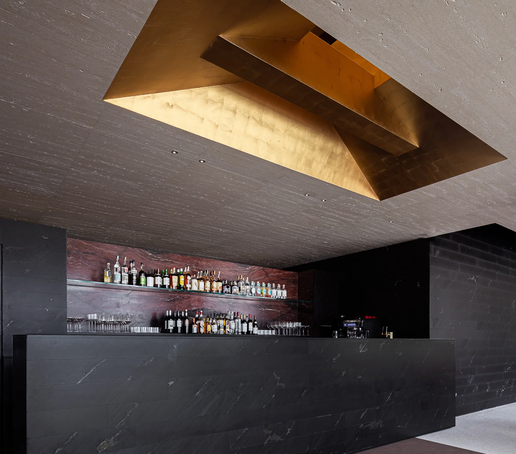 A modern black stone bar features a matching stone back wall, a dark red veined backsplash with glass shelves, and a gold-leaf recessed ceiling.