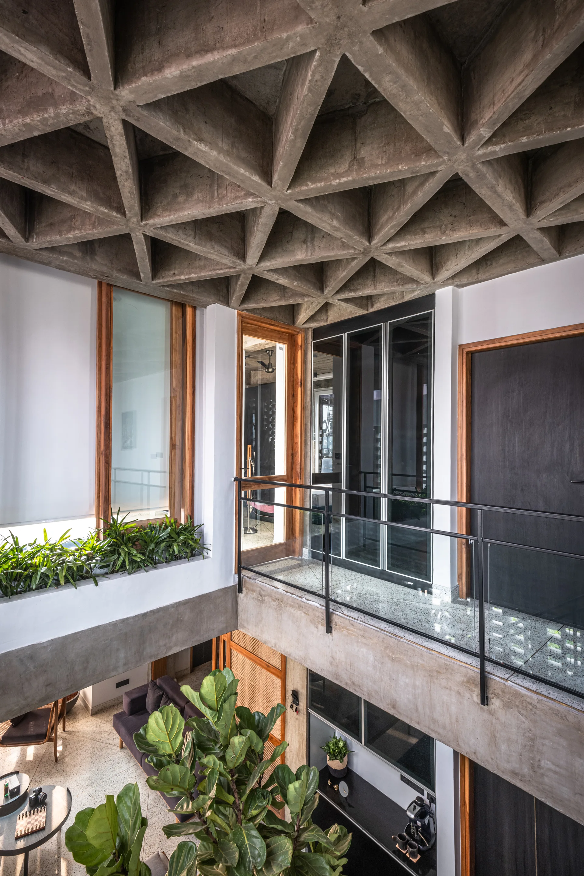 An interior mezzanine landing featuring an exposed concrete waffle ceiling, glass and metal railings, white walls with wooden doors, and green plants.