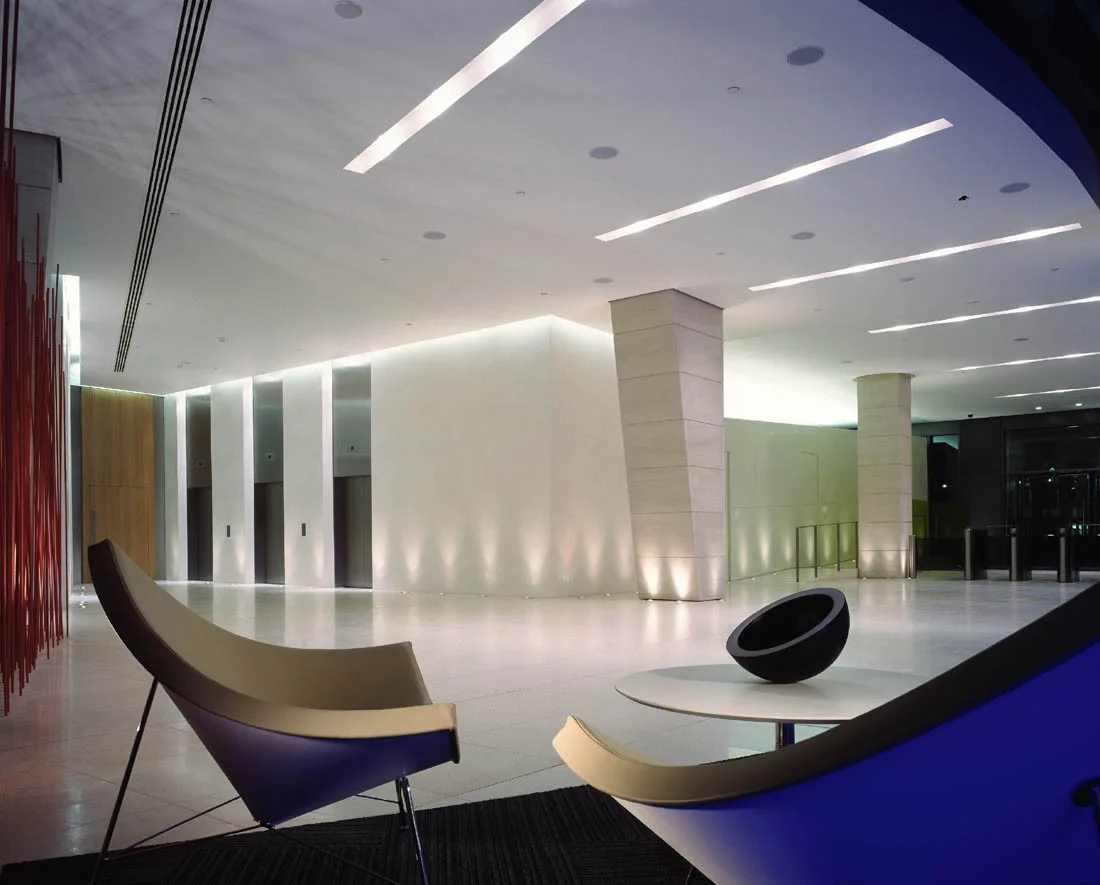 A spacious minimalist lobby with curved armchairs in the foreground, polished floors, massive stone columns, and recessed linear lighting.