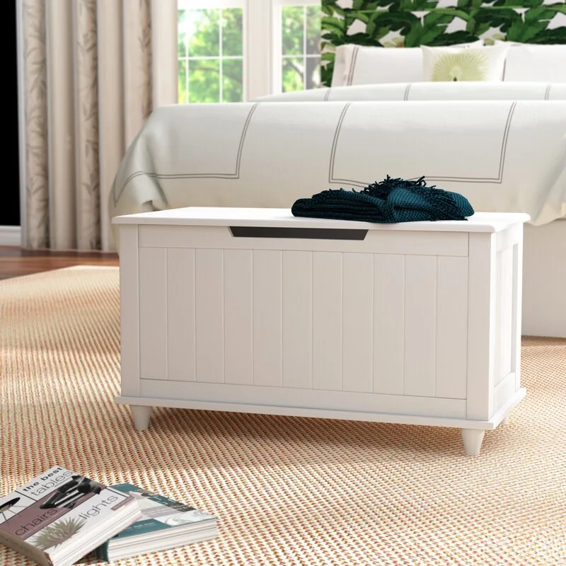 A white wooden storage chest with vertical slats sits on a textured tan rug at the foot of a bed with white linens.