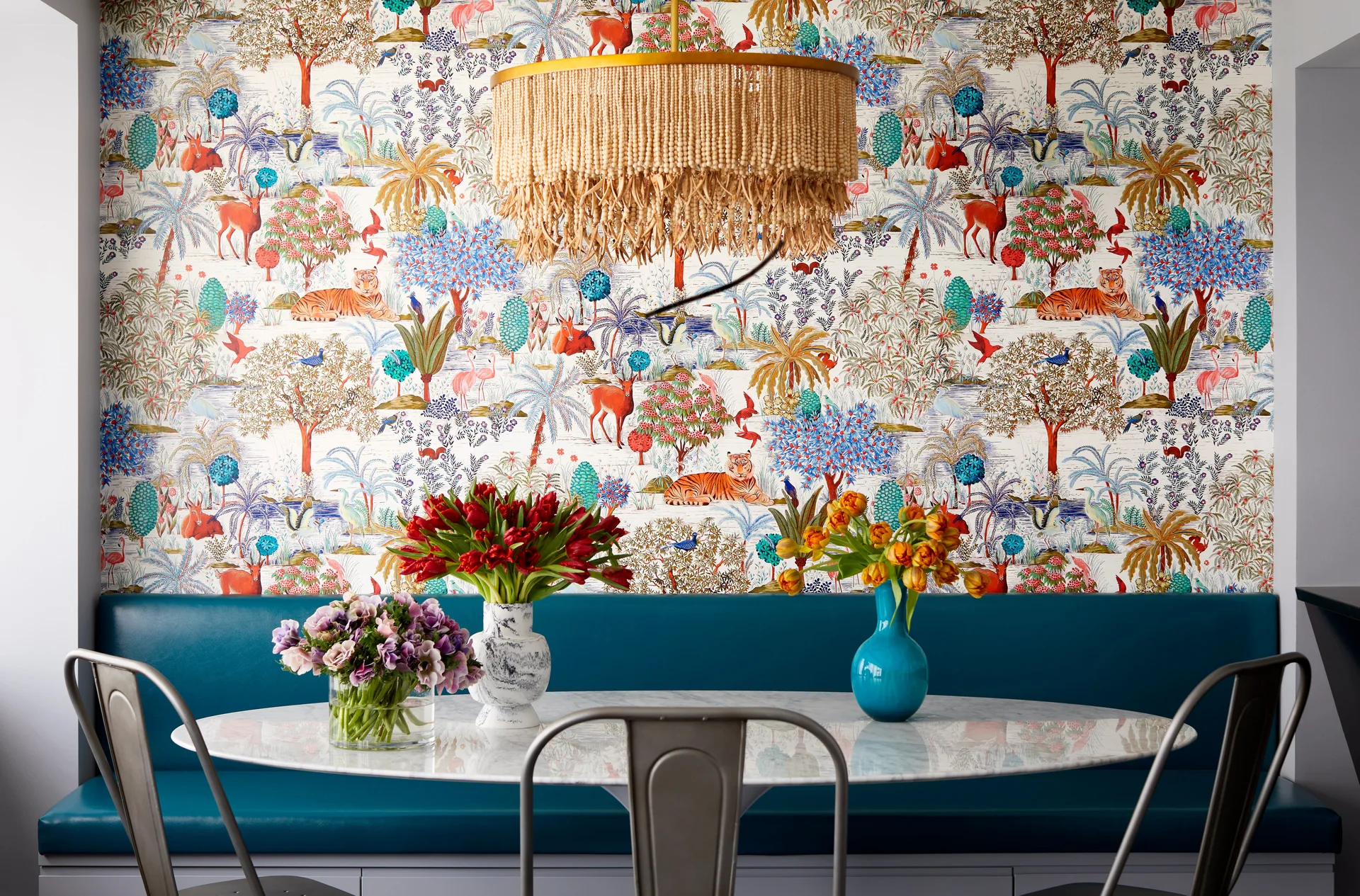 A dining nook features a teal upholstered banquette, a round marble-topped table, and vibrant jungle-themed wallpaper under a beaded pendant light.