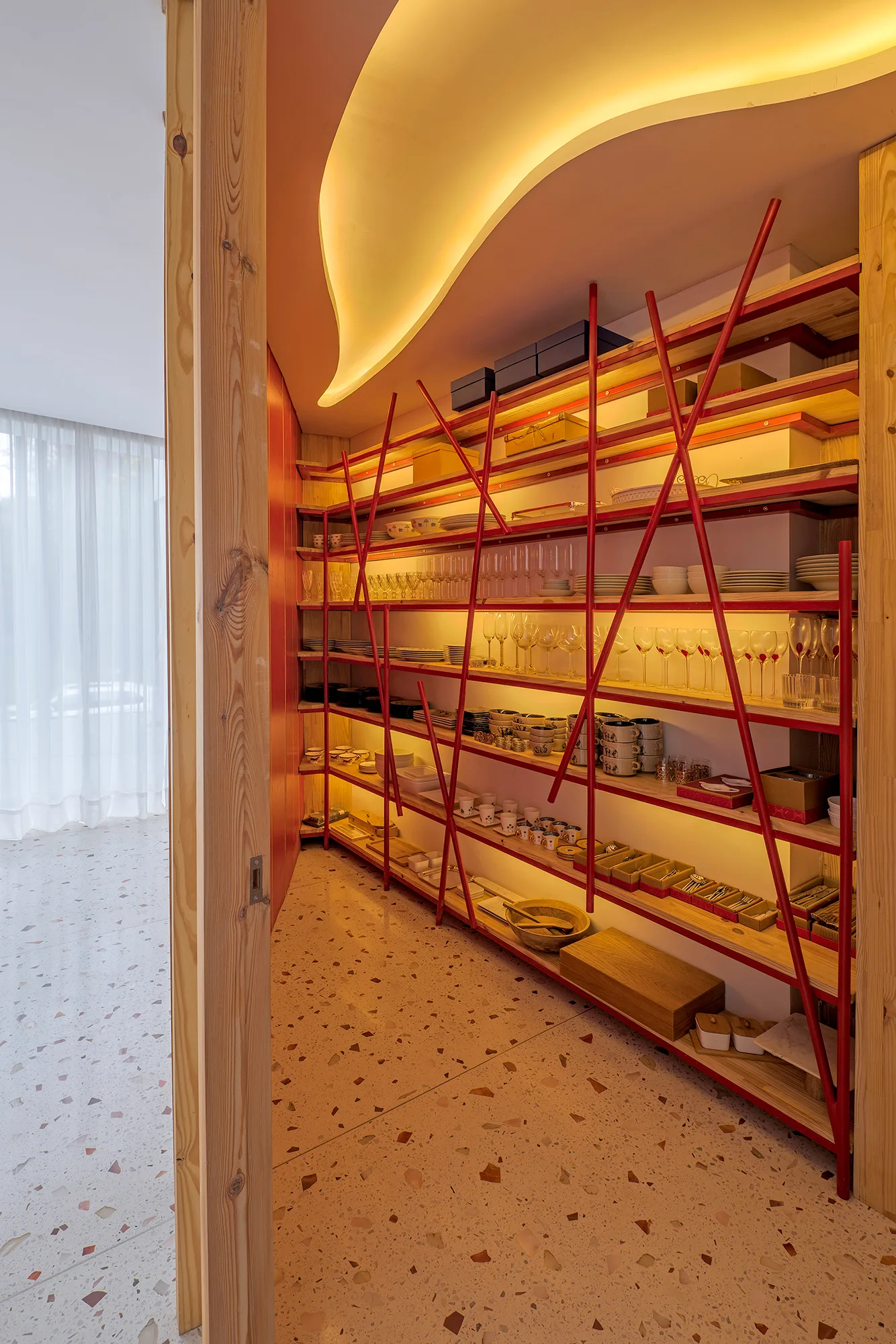 Open wooden pantry shelving features striking red metal cross bracing, illuminated by built-in warm lighting over a speckled terrazzo floor.