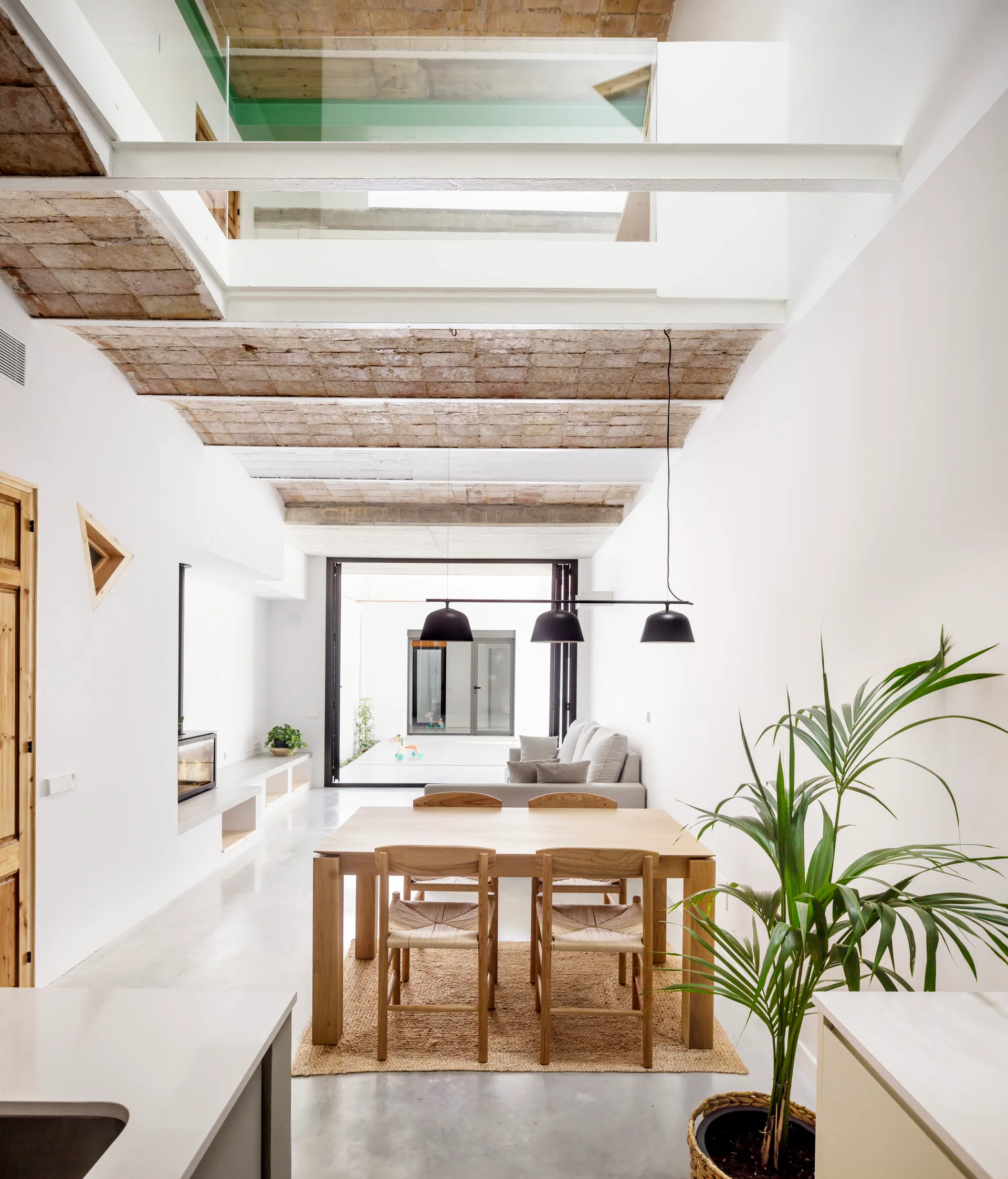 A modern open dining room featuring a light wood dining set under a vaulted exposed brick ceiling, with a concrete floor and white walls.