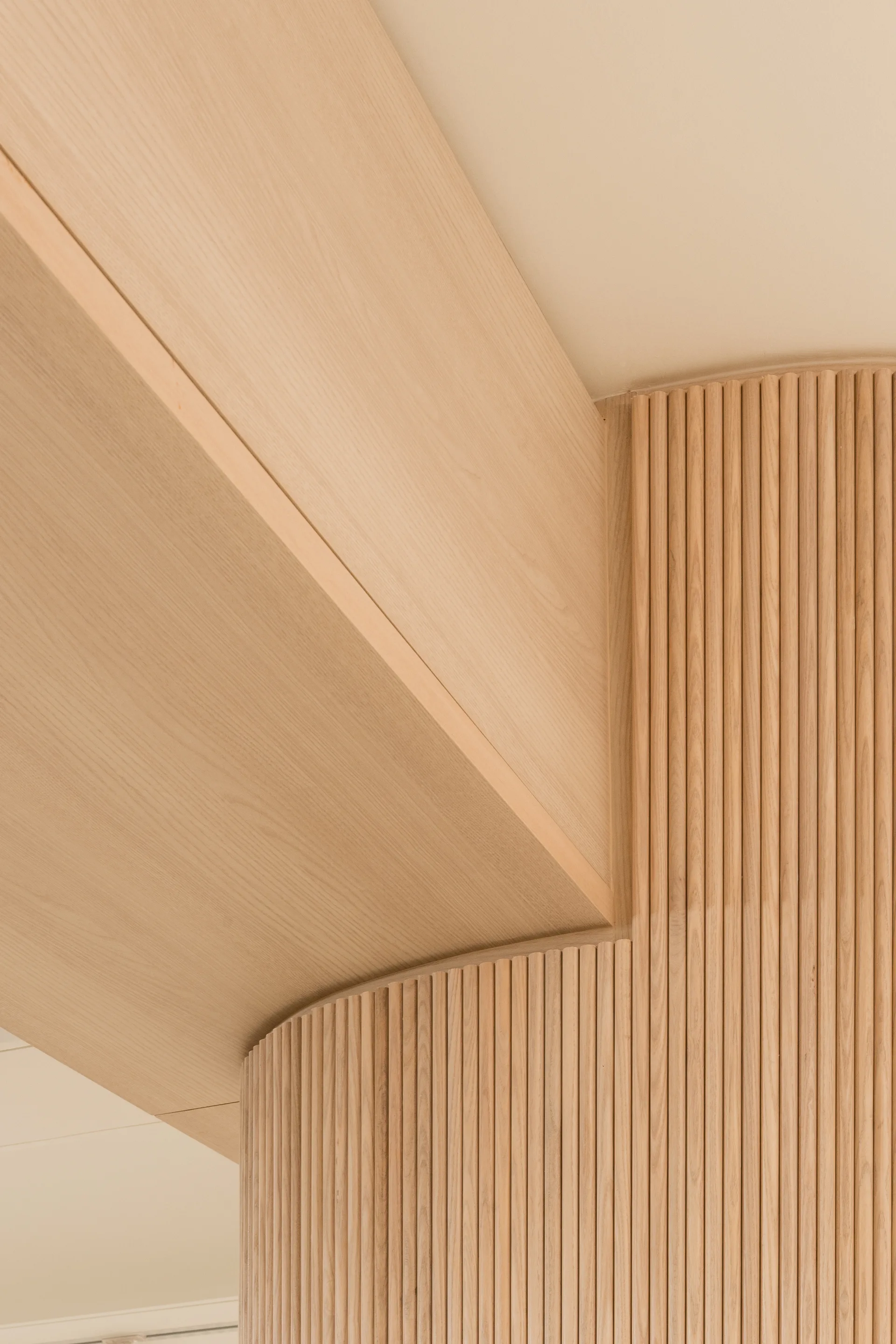 Close-up of minimalist architectural joinery featuring curved light-toned fluted wood wall paneling and a smooth overhead wood soffit.