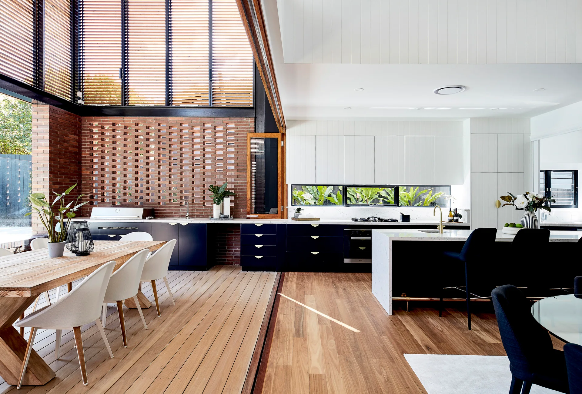 A modern kitchen with navy blue base cabinetry, white countertops, an open dining area on a wood deck, and an exposed brick wall.