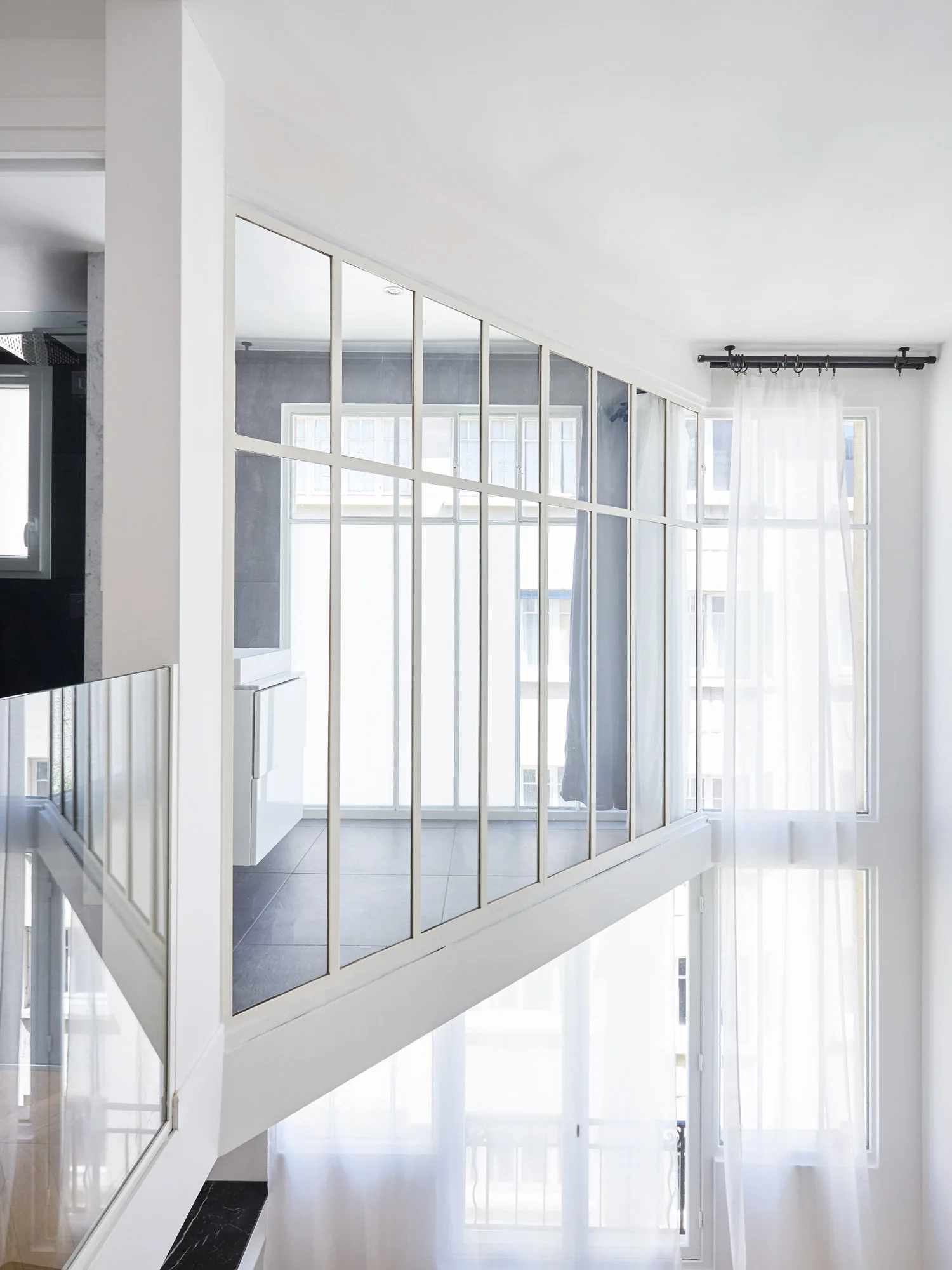 An interior view of a white mezzanine level featuring a large grid-patterned glass partition and tall windows with sheer curtains.