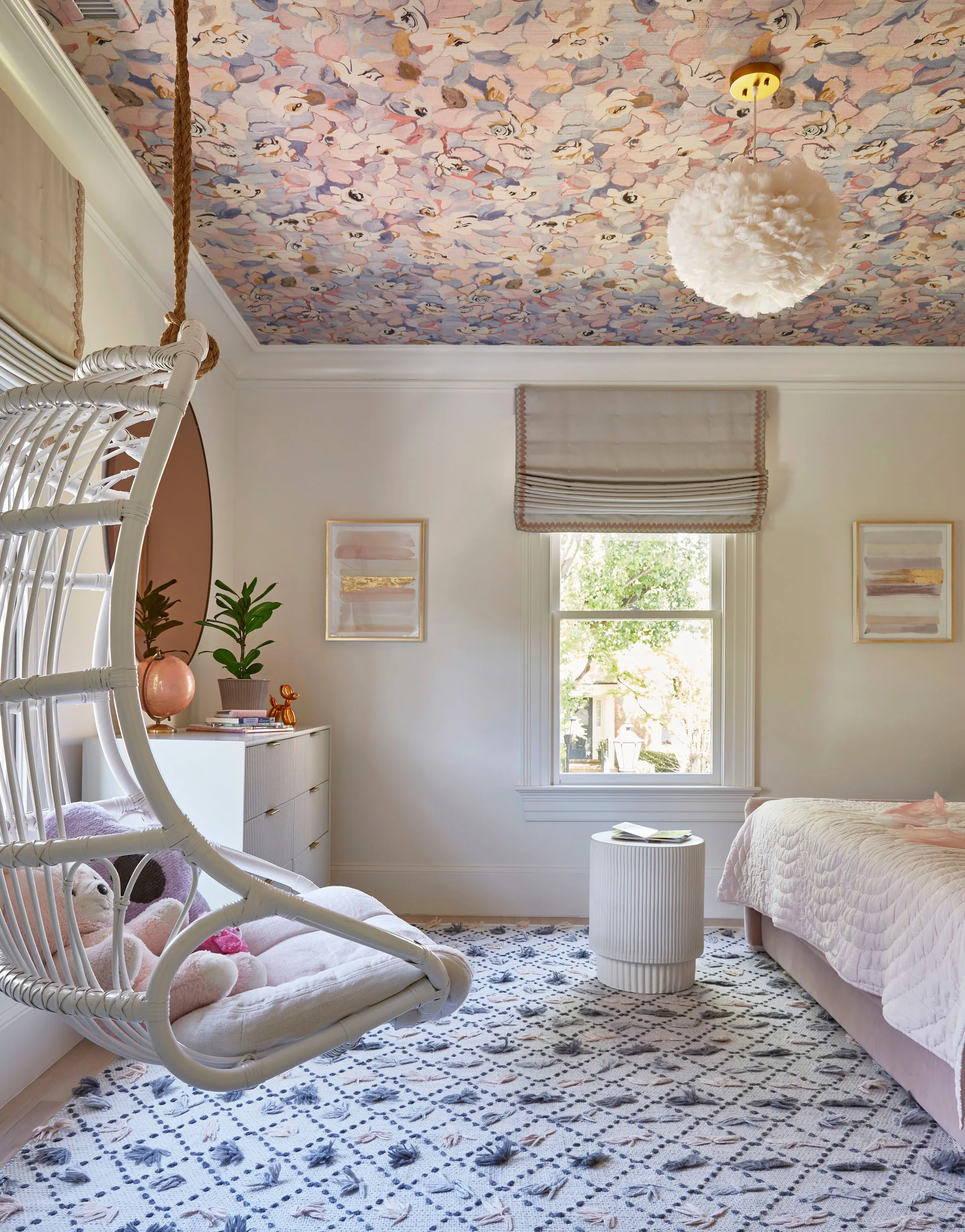 A bright bedroom features a white hanging wicker chair, a bed with pink bedding, a textured rug, and colorful floral wallpaper on the ceiling.