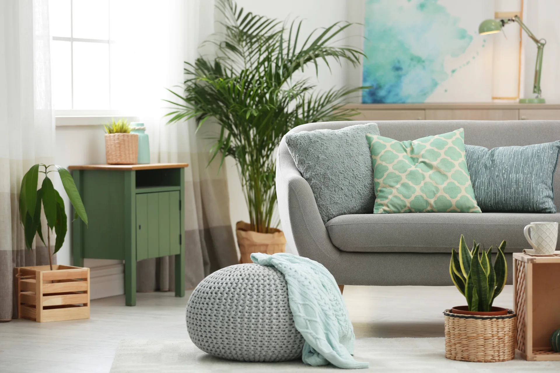 A bright living room features a grey sofa with green patterned pillows, a green side table, a knitted pouf, and potted houseplants.