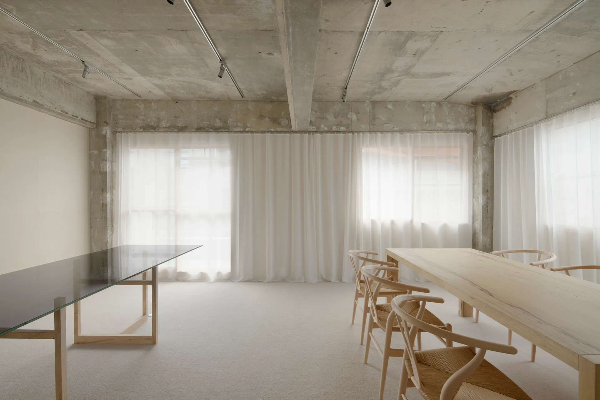 A minimalist dining room features a long wooden table with wishbone chairs beneath an exposed concrete ceiling with floor-to-ceiling sheer curtains.
