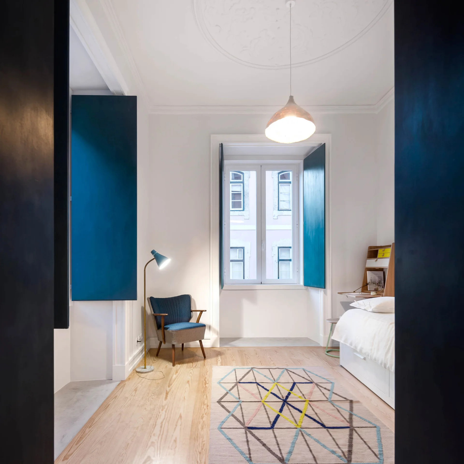 A modern white bedroom features bright blue folding window shutters, a blue armchair, a geometric patterned rug, and a central pendant light.