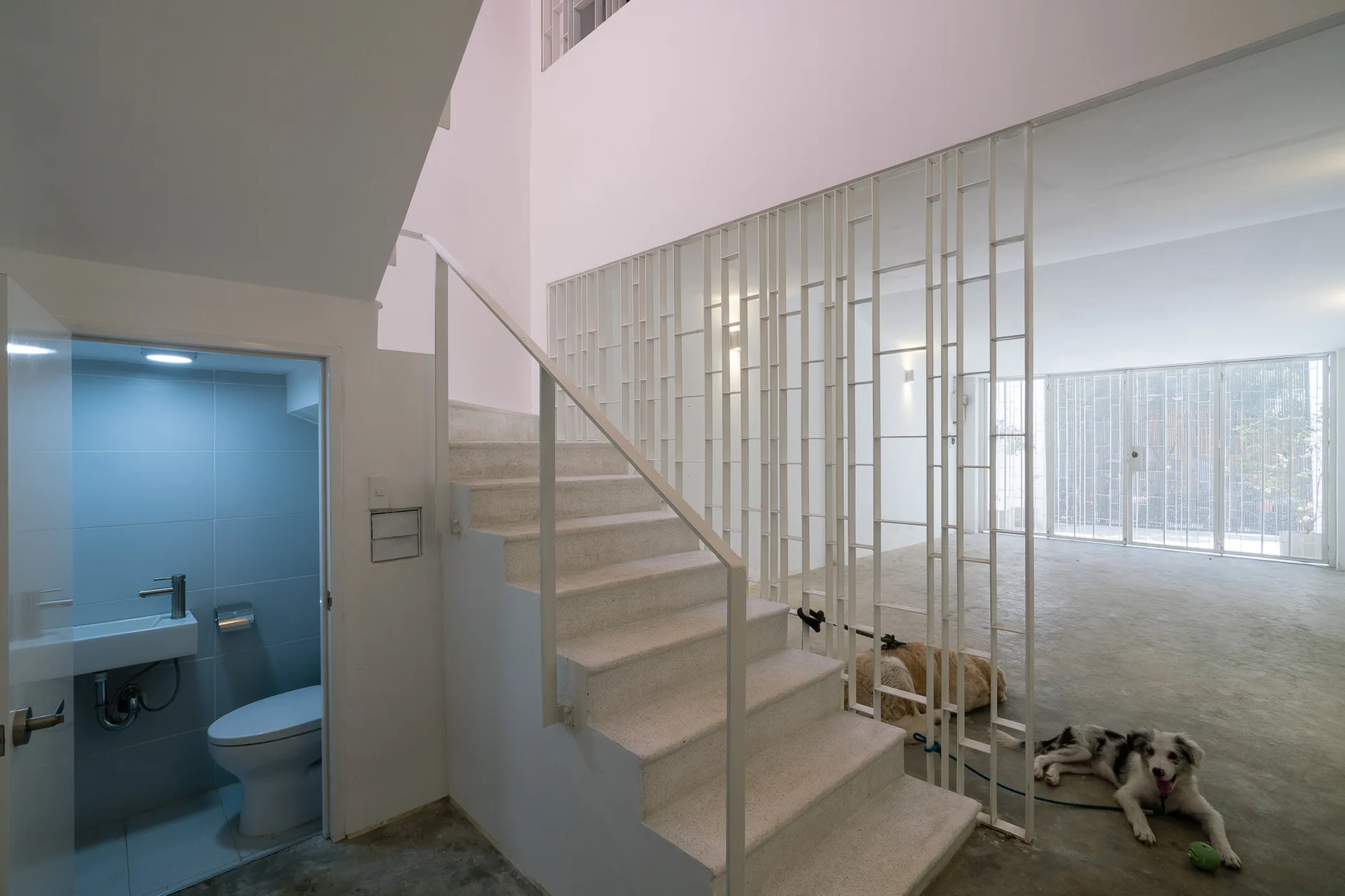 A minimalist white staircase next to a geometric metal screen room divider and an open door showing a small powder room with a polished concrete floor.