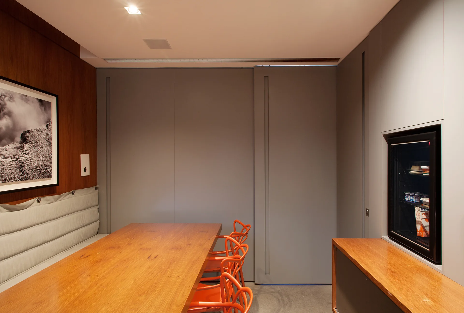A wooden dining table is paired with bright orange chairs, a grey tufted wall bench, and minimalist grey storage cabinets.