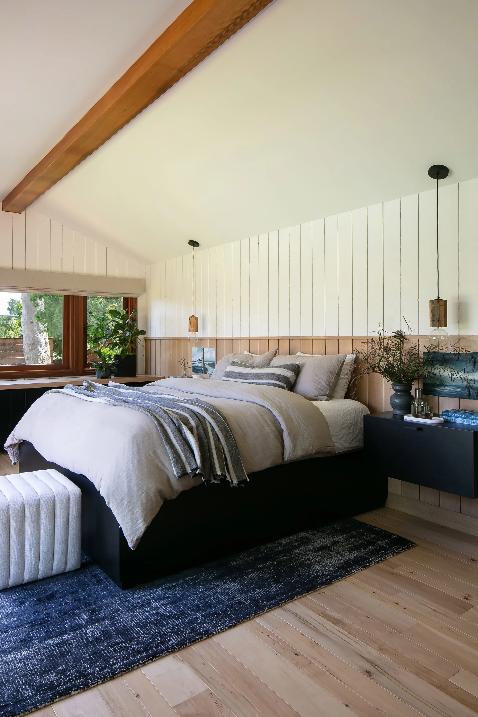 A modern bedroom features a platform bed with grey linens, split wood-paneled walls, a ceiling beam, and a floating black nightstand.
