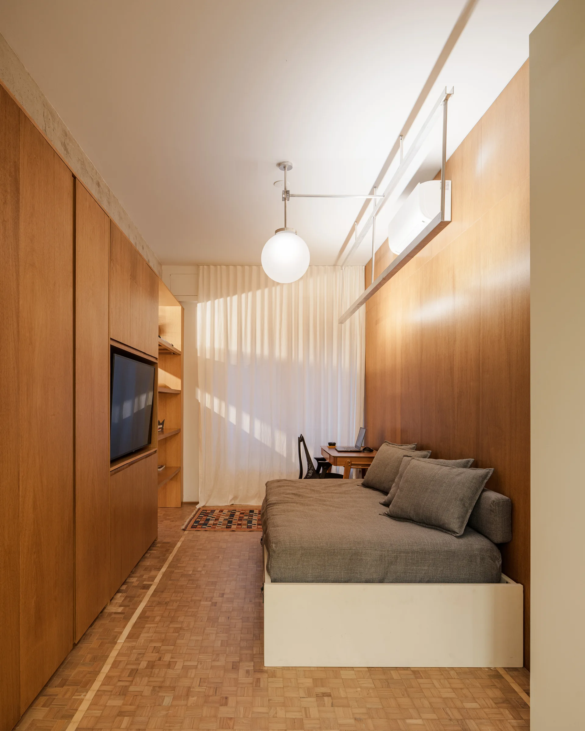A narrow modern bedroom features warm wood-paneled walls, built-in storage cabinets, a grey daybed, and parquet wood flooring under a globe pendant light.