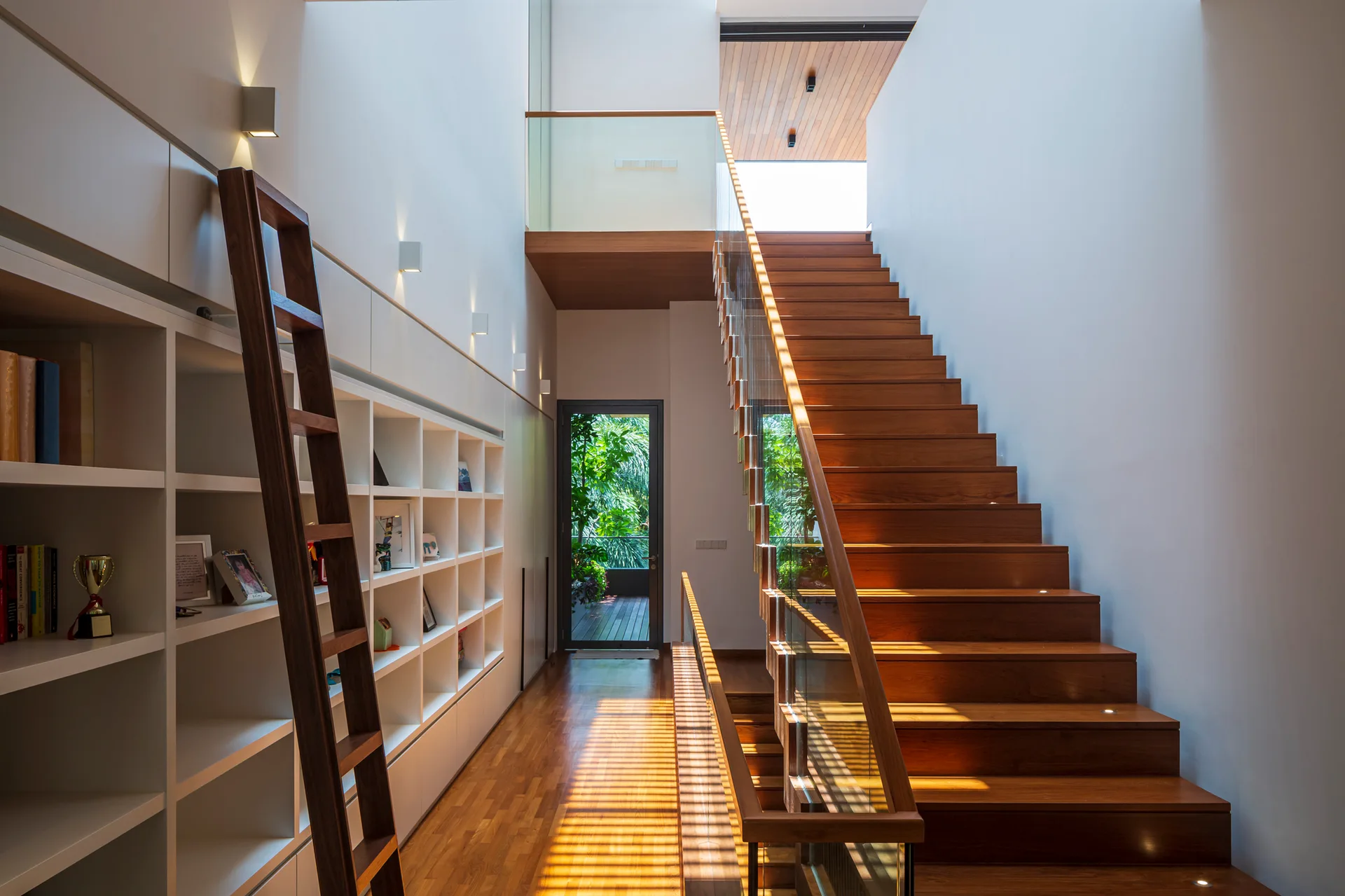 A modern hallway featuring a warm wooden staircase with a glass railing opposite a built-in white bookshelf and matching wood ladder.