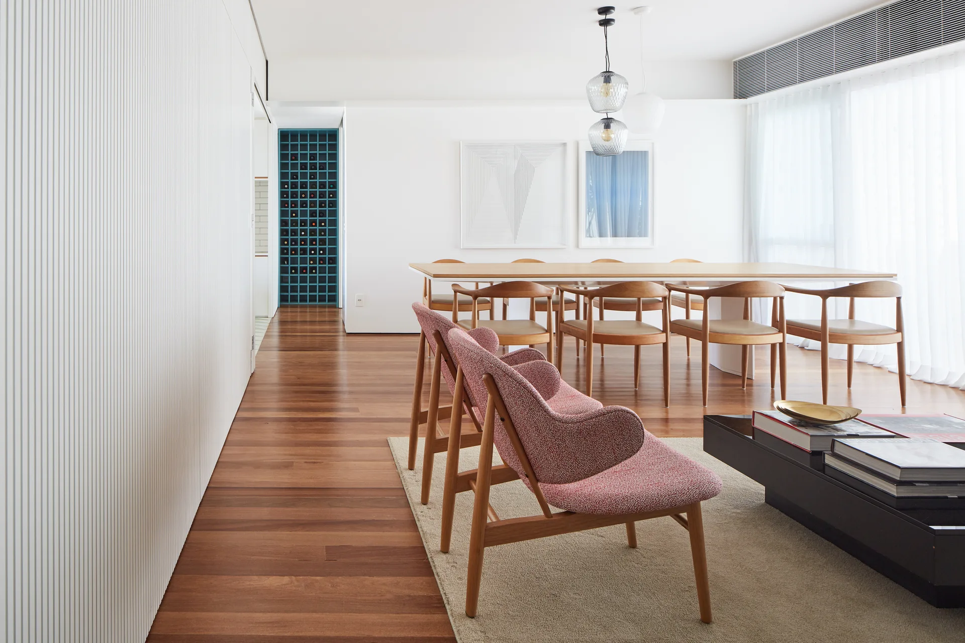 A modern dining room features a light wood table with curved chairs, white slatted wall paneling, and two pink armchairs in the foreground.