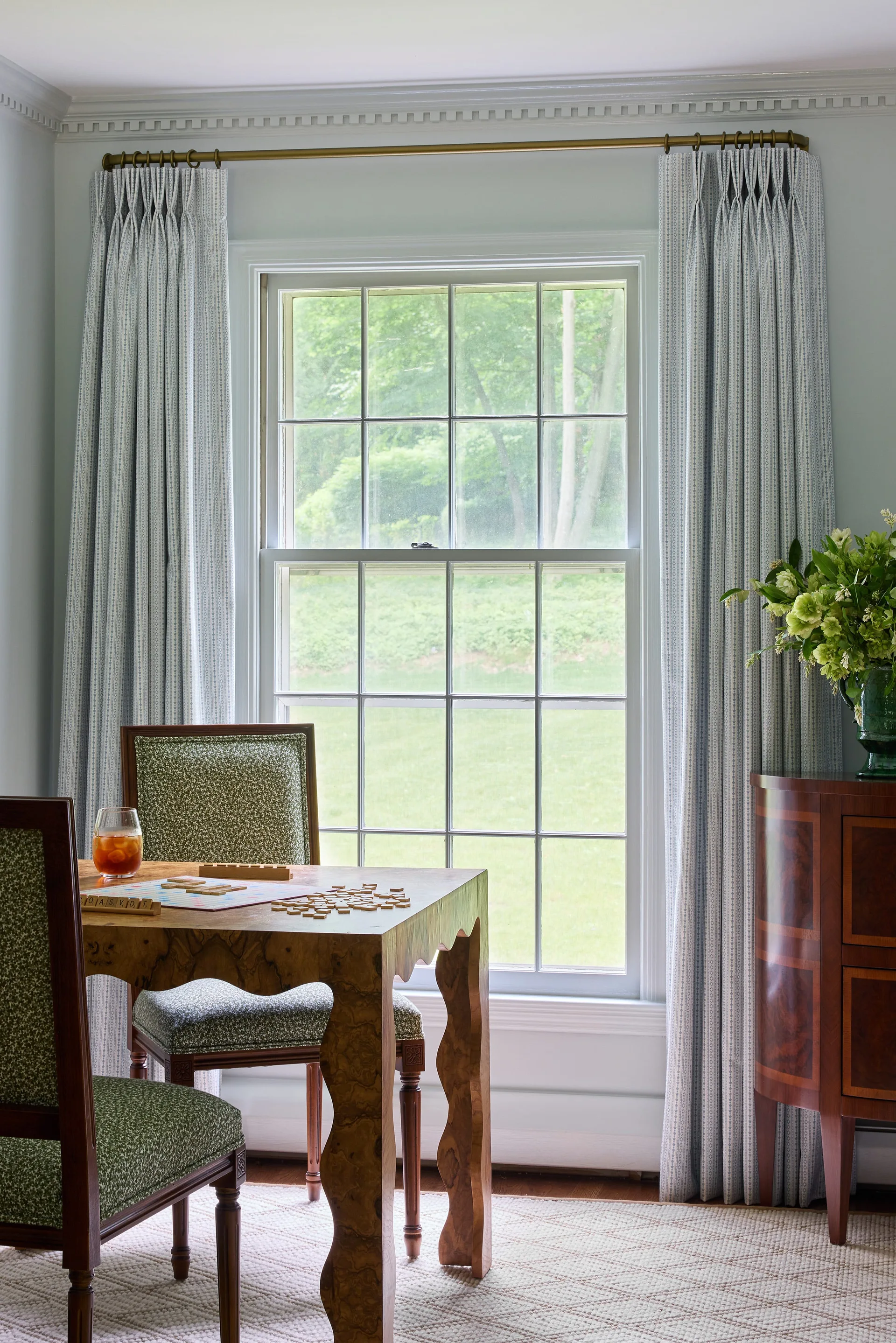 A burl wood game table with green patterned chairs sits before a window flanked by light blue striped drapes next to a curved wood sideboard.