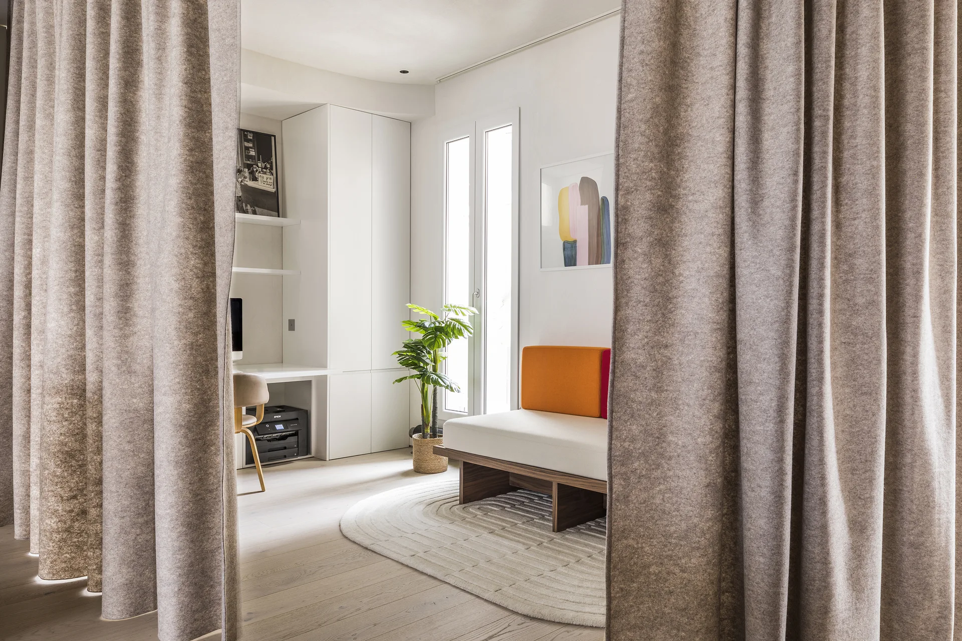 Heavy beige curtains frame a bright room containing a white built-in desk, a low wooden daybed with an orange backrest, and an oval rug.
