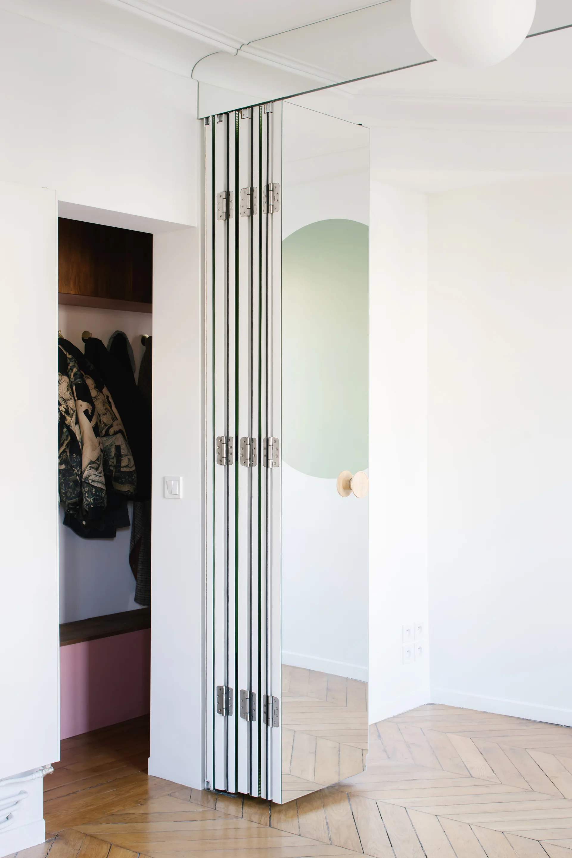 A mirrored bifold closet door is partially folded open, revealing a wardrobe interior, set against white walls and light herringbone wood floors.