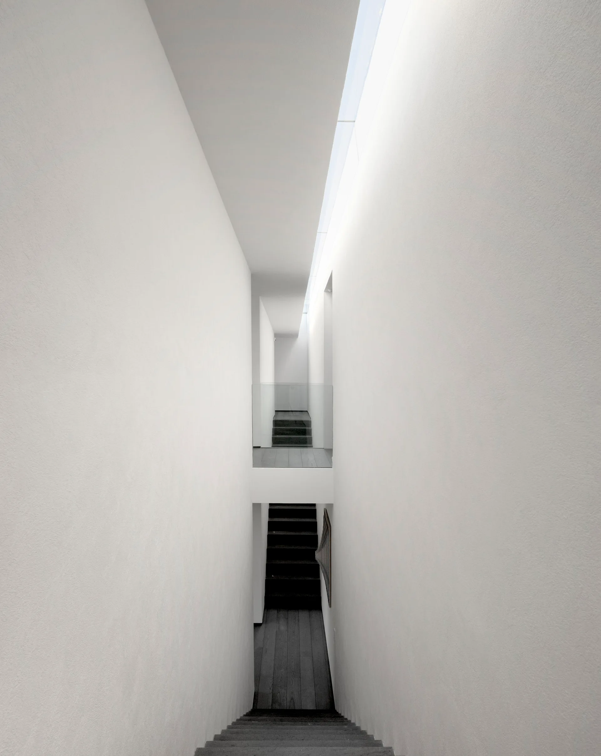 A high-angle view of a narrow minimalist staircase with dark wood treads, tall smooth white walls, and a long linear skylight overhead.