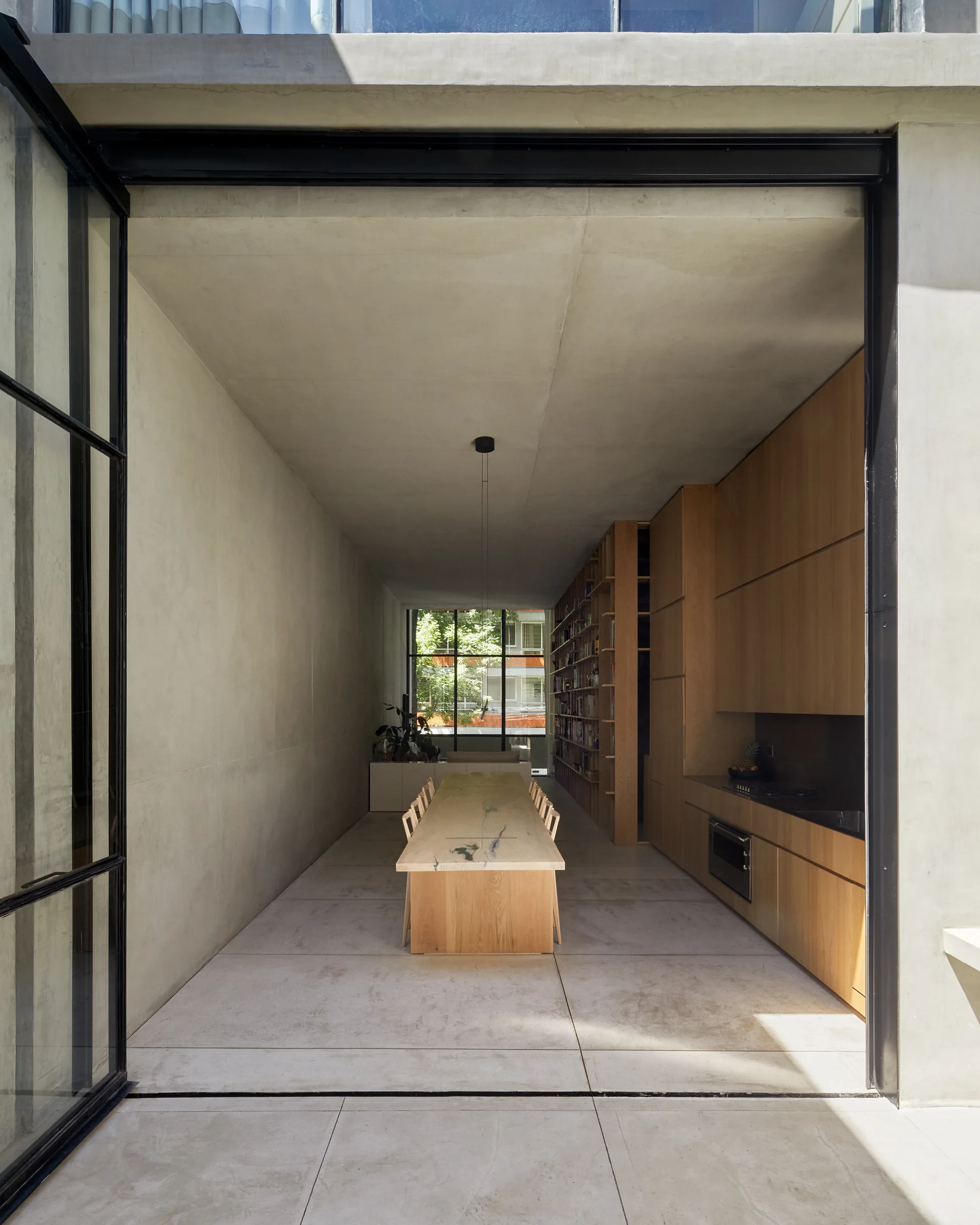 A long wooden dining table sits in a minimalist room with smooth concrete walls, built-in timber kitchen cabinetry, and a large black-framed glass entryway.