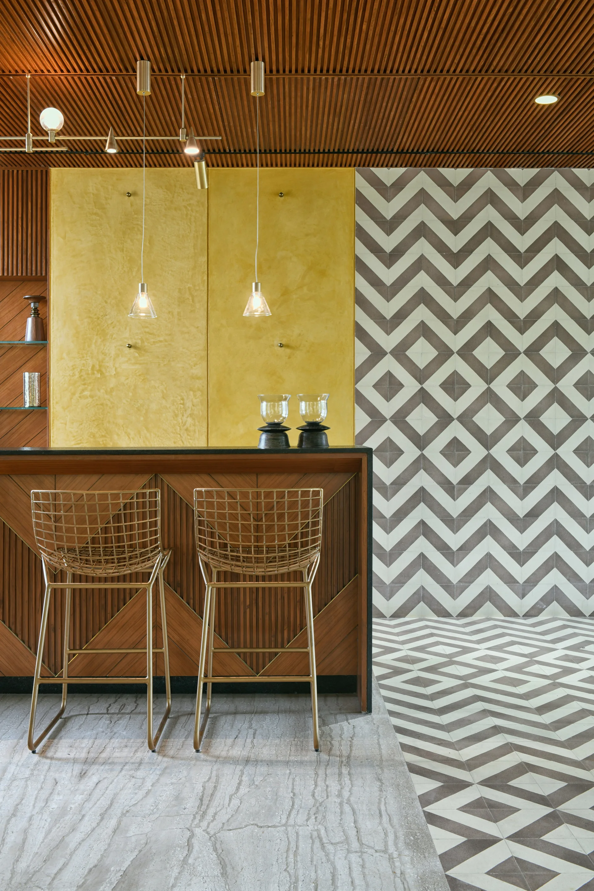 A modern home bar features two gold wire stools at a wooden counter, a mustard yellow accent wall, and geometric chevron patterned floor and wall tiles.