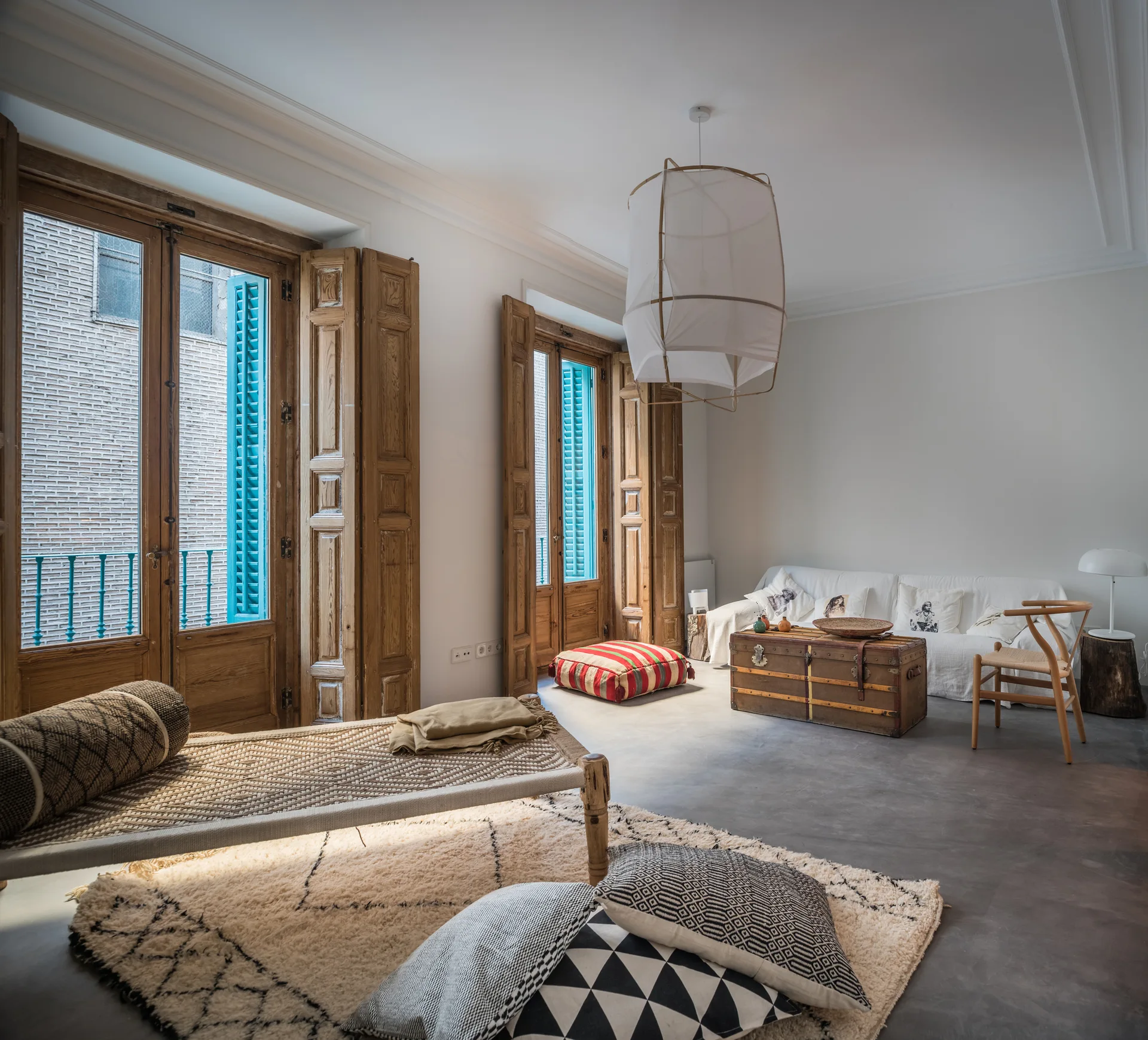A rustic living room features a woven daybed, a vintage trunk coffee table, concrete floors, and large wooden windows with bright blue shutters.