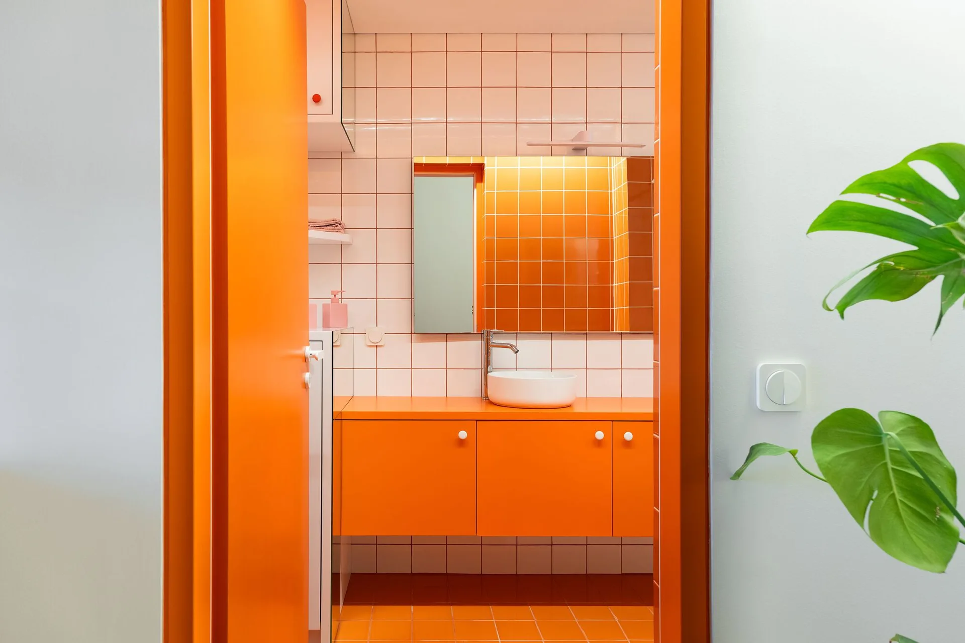 A view through an orange doorway into a bathroom featuring a bright orange vanity cabinet, white vessel sink, and white grid-patterned tile walls.