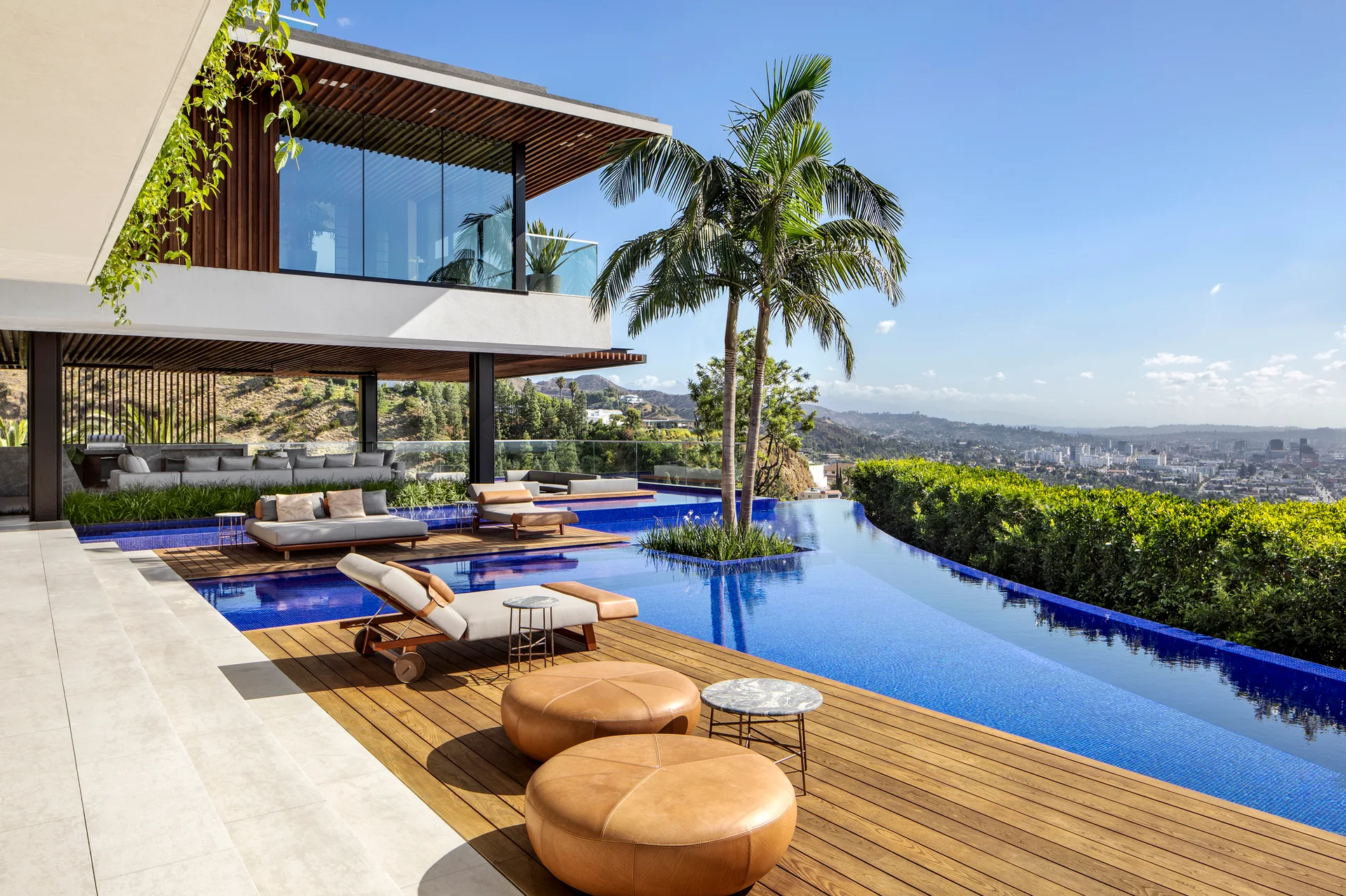 A modern outdoor pool patio features a blue infinity pool, wood decking, lounge chairs, and circular tan ottomans overlooking a hillside view.