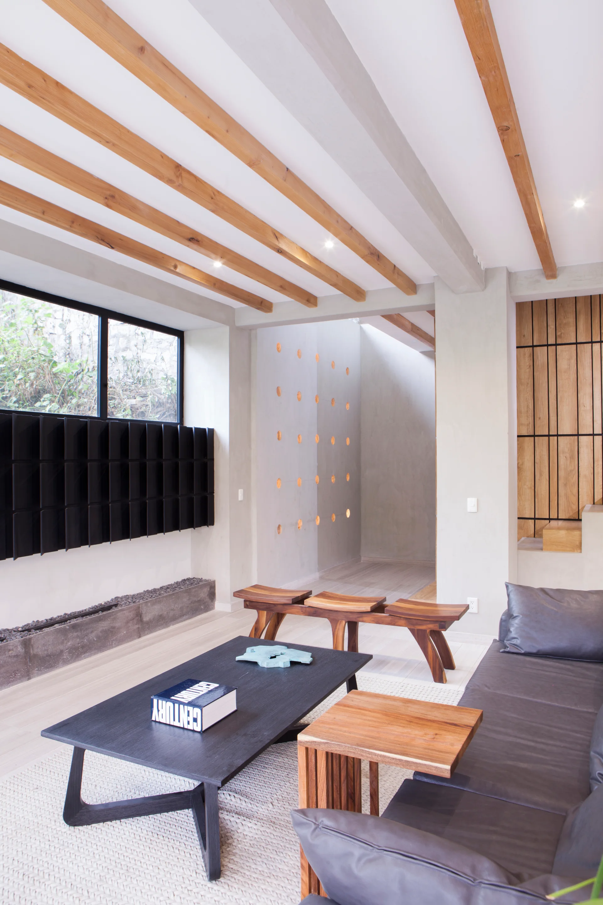 A modern living room features a dark gray sofa, a low black coffee table, exposed wood ceiling beams, and smooth concrete walls.