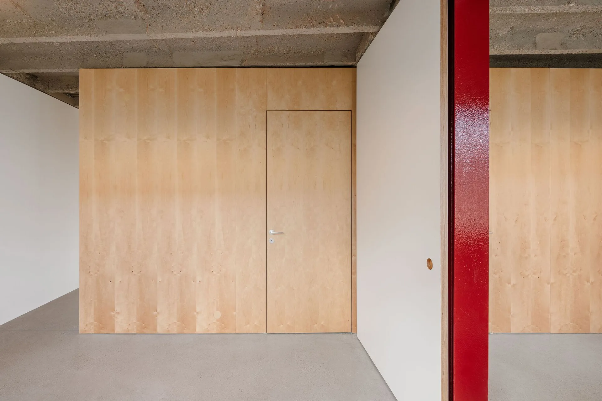 A minimalist interior features light plywood wall panels, a matching flush door, a raw concrete ceiling, and a bright glossy red structural column.