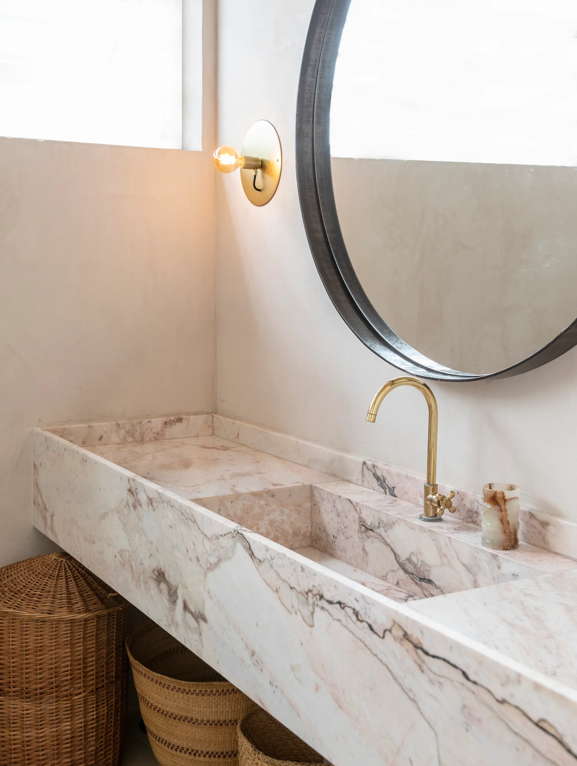 A modern wall-mounted pink marble trough sink with brass fixtures, a round mirror, and woven storage baskets underneath.