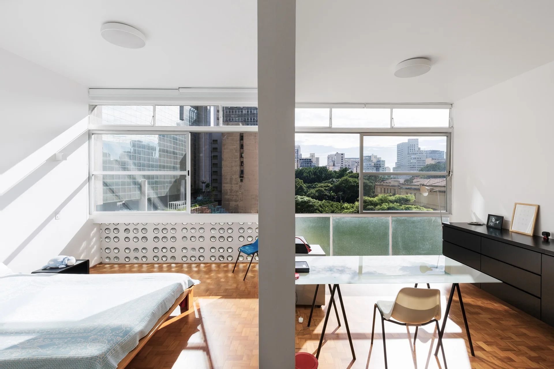 A bright minimalist bedroom and study area separated by a central white column, featuring parquet floors, a glass desk, and large windows.