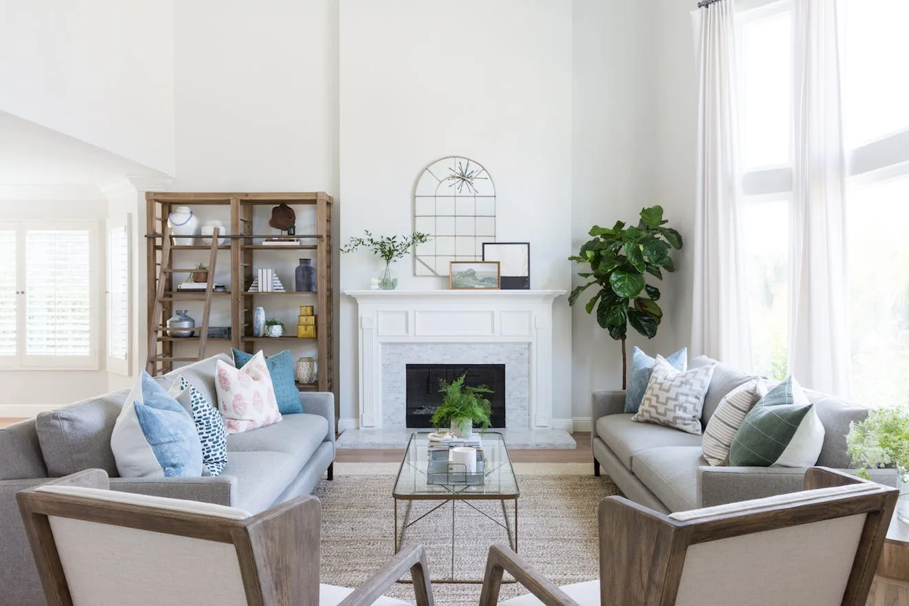 A bright living room features two gray sofas facing each other, a central white fireplace, a tall wooden bookcase, and a glass coffee table on a woven rug.