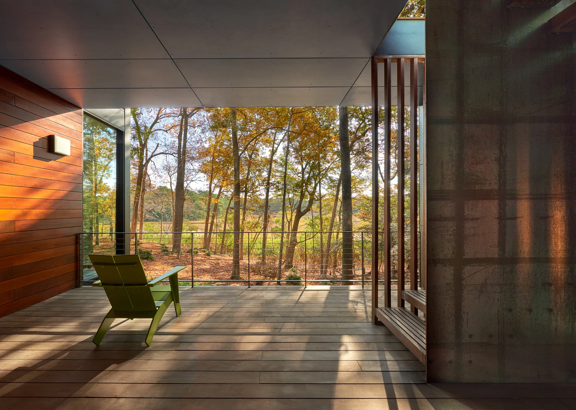 A covered wooden deck features a lime green Adirondack chair, cable railing, and a view of autumn forest trees.