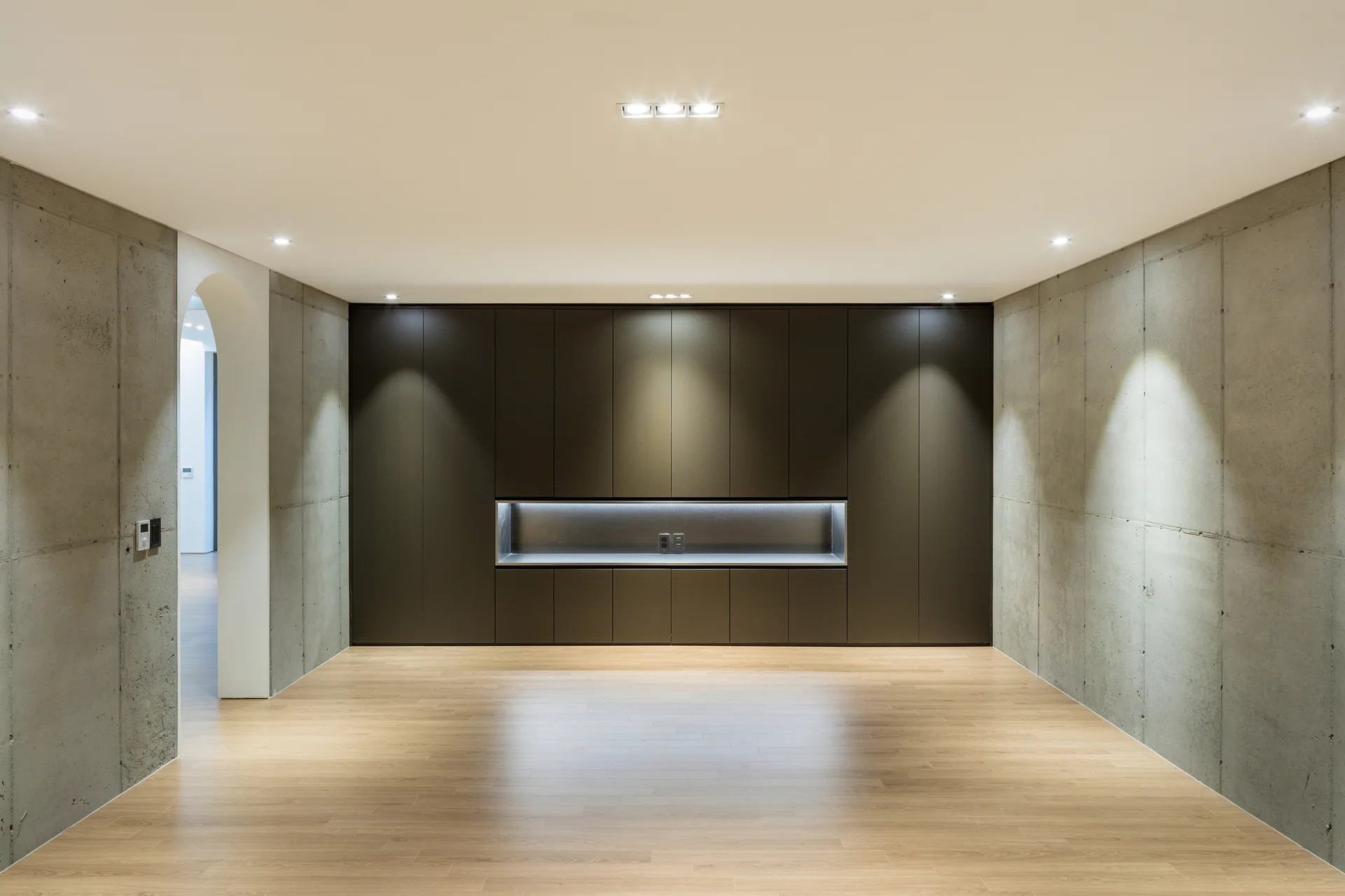 A minimalist room featuring raw concrete walls, light wood flooring, an arched doorway, and a large dark built-in cabinet with a lighted recess.