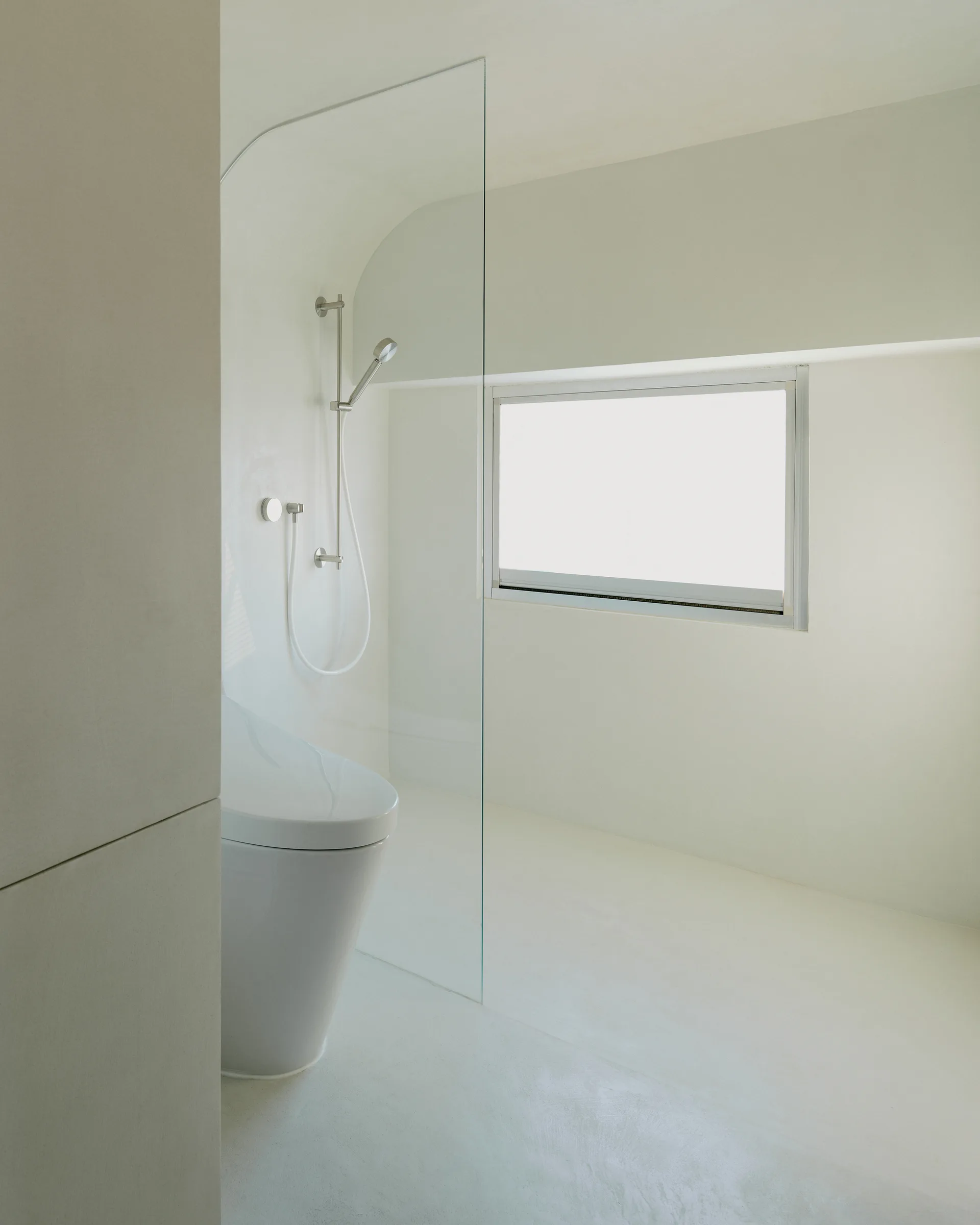 A minimalist all-white bathroom featuring a seamless plaster finish, a frameless glass shower partition, a modern toilet, and a recessed window.