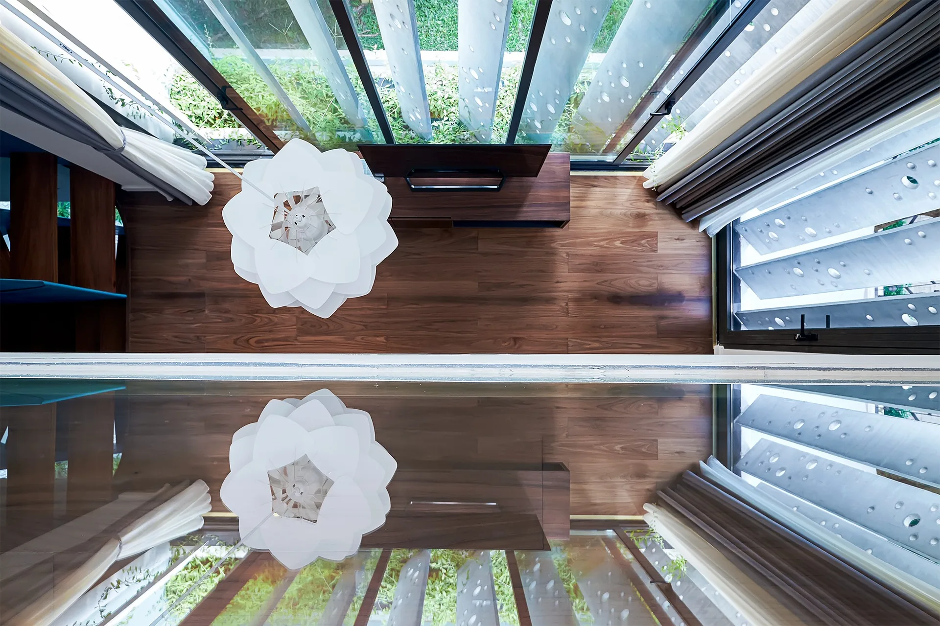 Top-down view of a modern room with dark wood flooring, a white floral pendant light, and a media console reflected in a large glass pane.