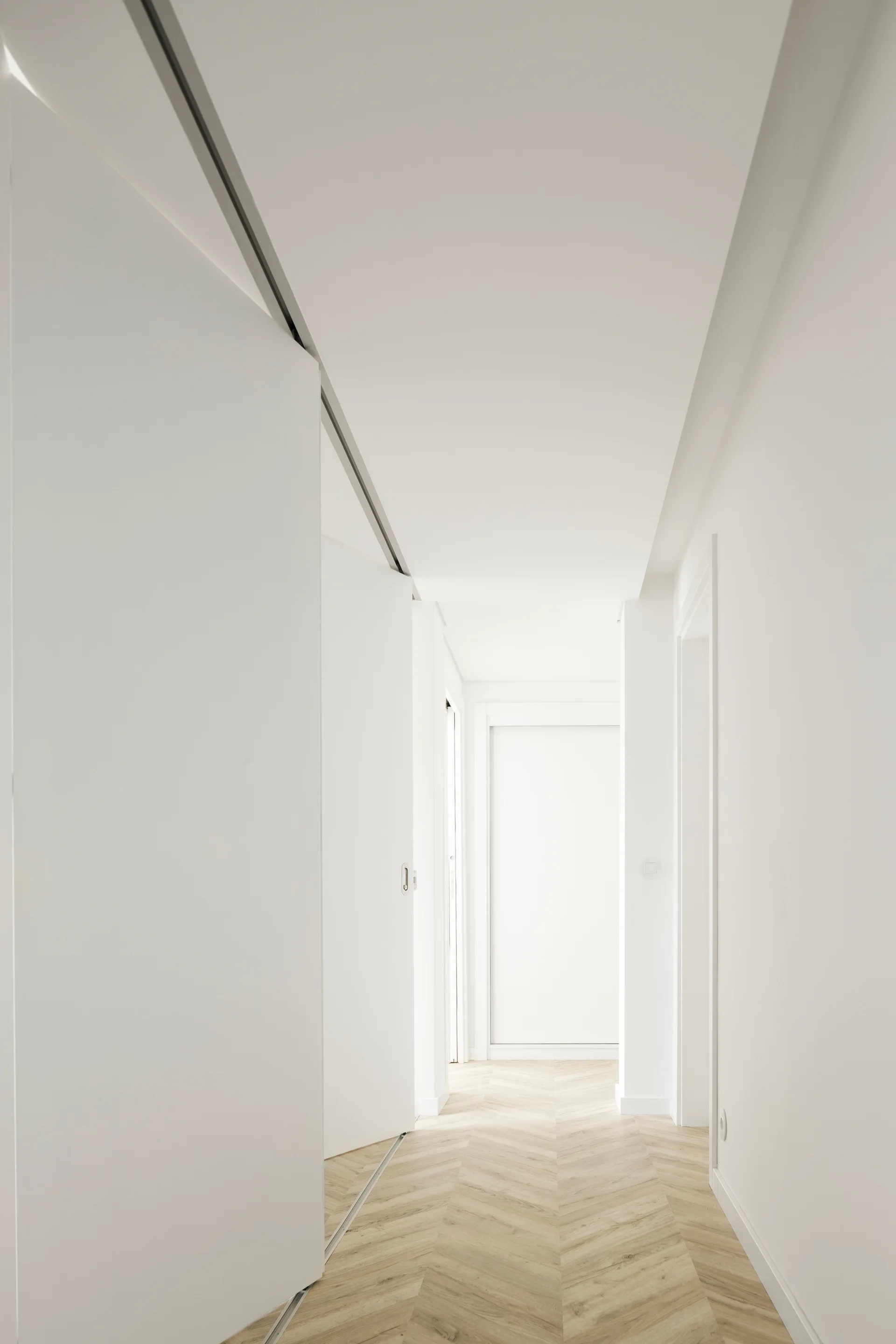 A bright minimalist white hallway features light wood herringbone parquet flooring and an integrated white sliding door.