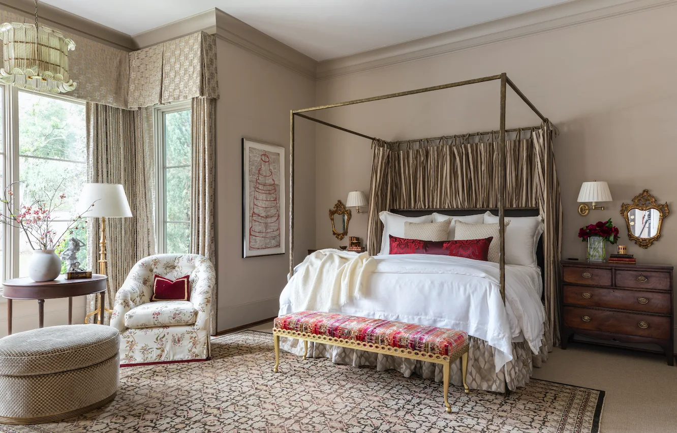 A traditional bedroom features a metal four-poster bed, an upholstered floral armchair by the window, and an ornate patterned rug.