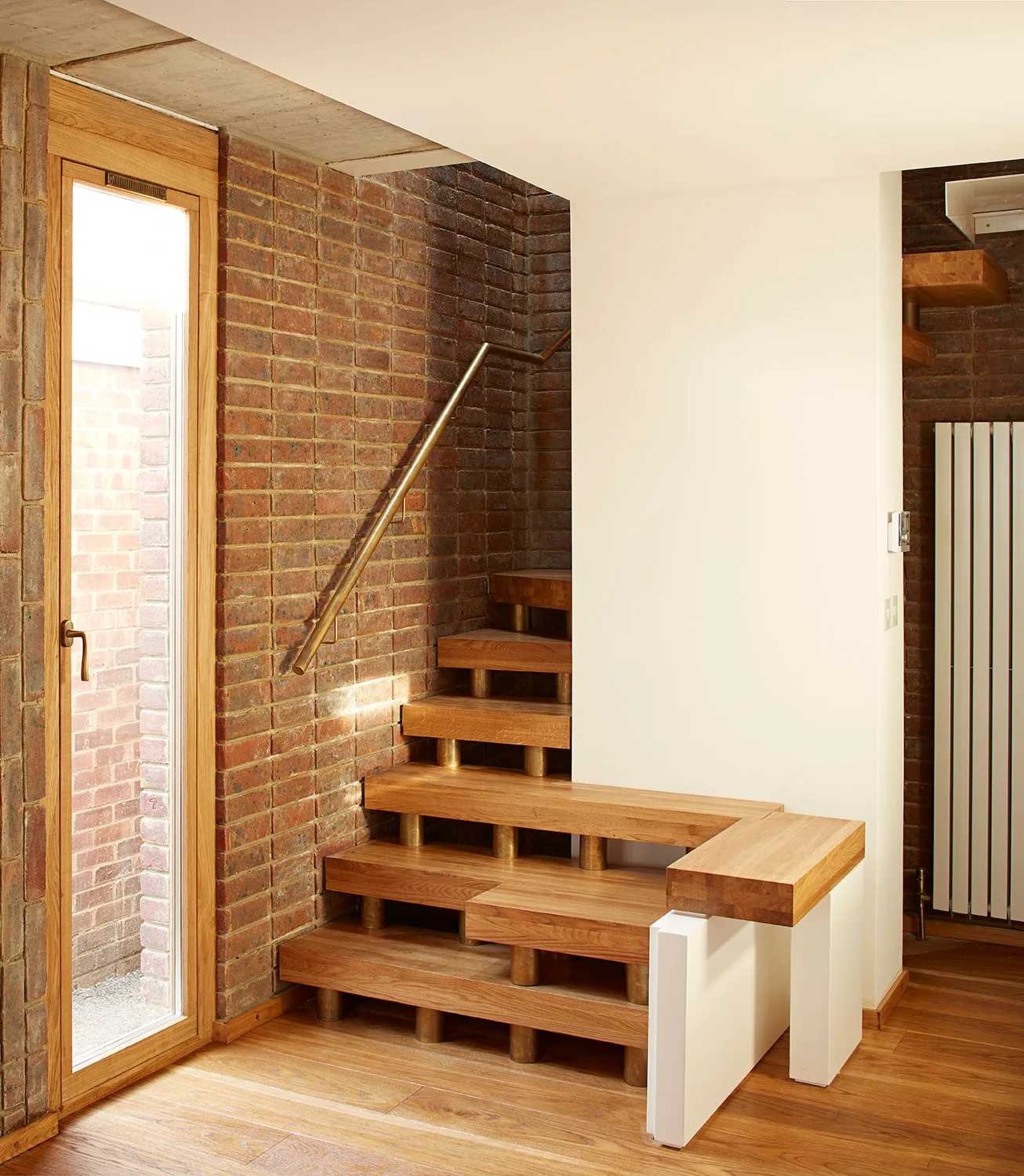 A wooden staircase with brass cylinder supports rises against an exposed red brick wall next to a glass-paned door.