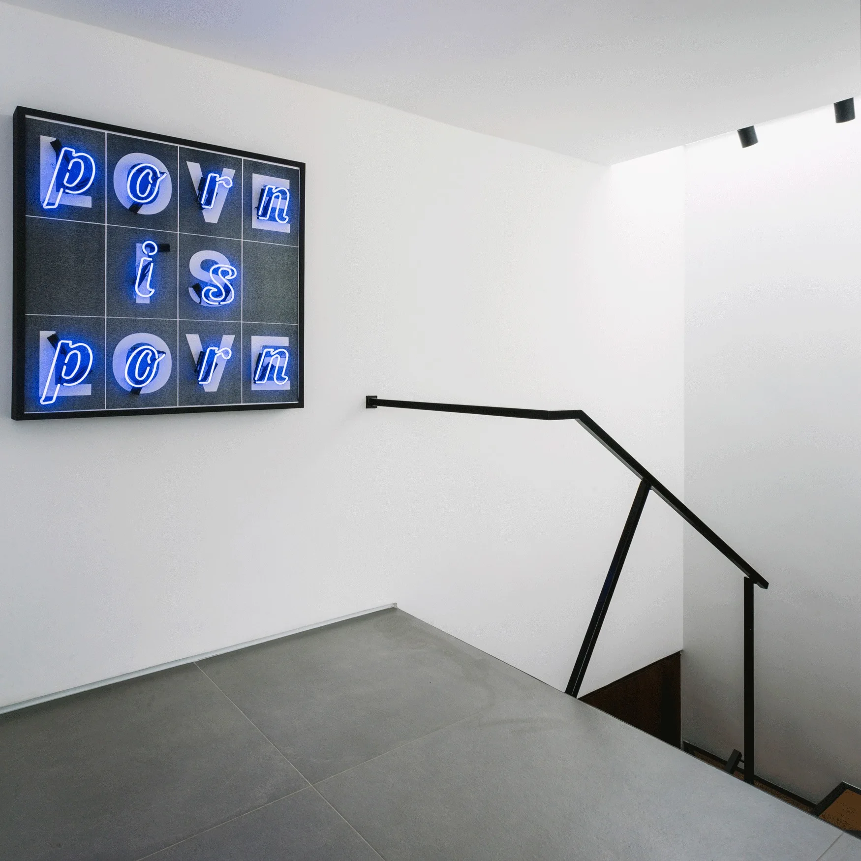 A minimalist staircase landing features grey tile flooring, white walls, a black metal handrail, and a framed blue neon wall art piece.
