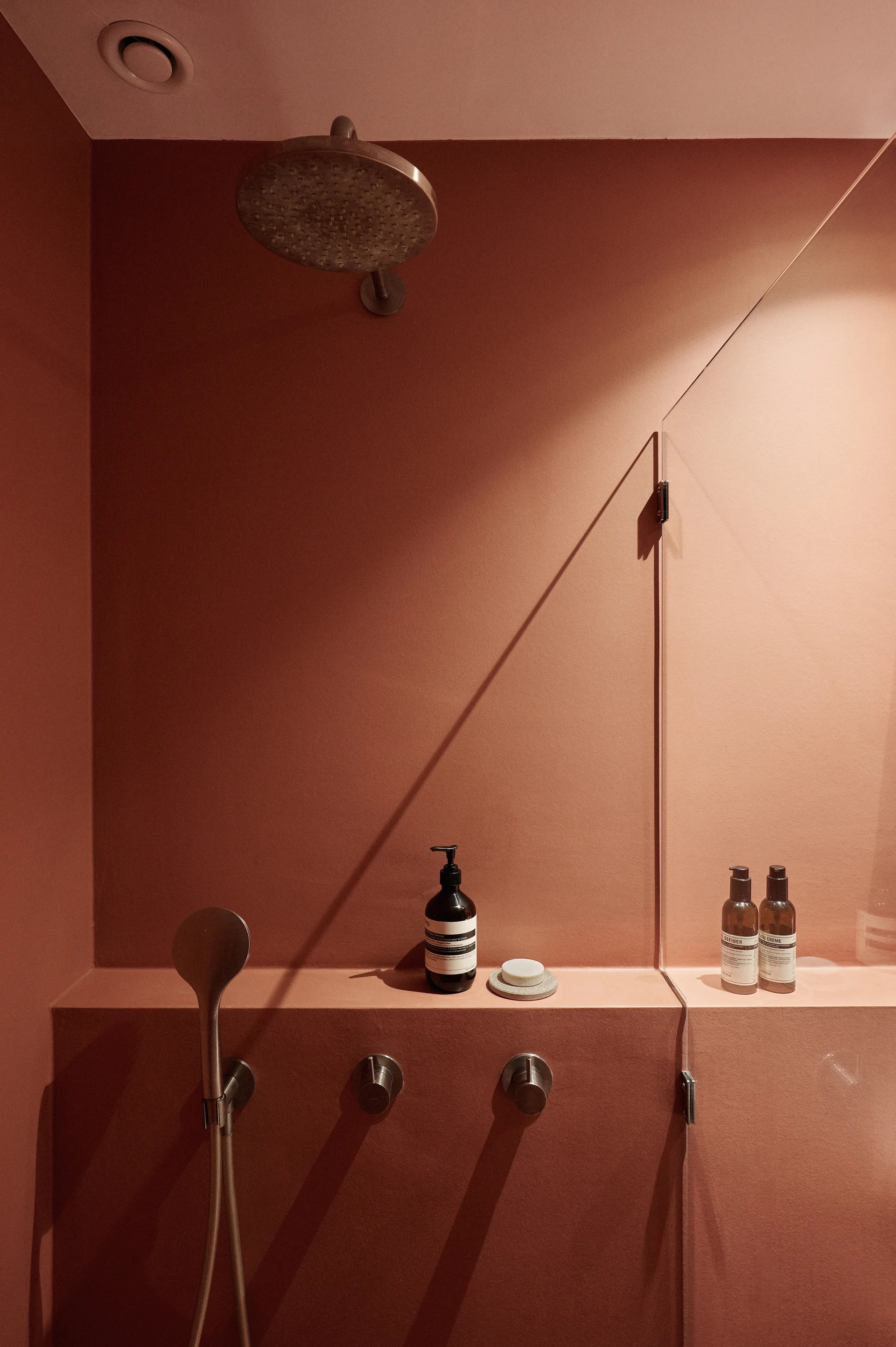 A minimalist terracotta shower features a built-in shelf ledge, brushed metal fixtures, and a glass partition panel.